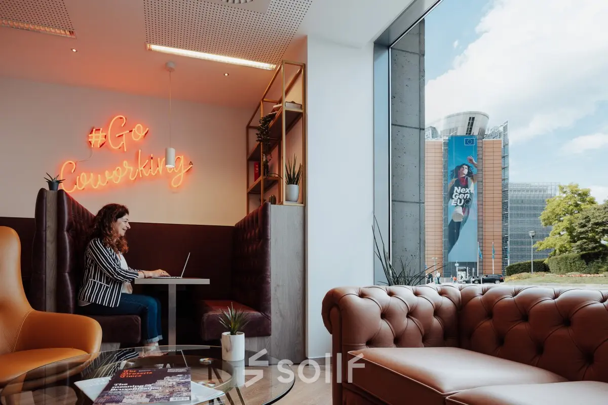 Moderne, gemeubileerde kantoor te huur op het Robert Schumanplein 2-4, Brussel Europese Wijk, met uitzicht op de straat en een persoon die aan een laptop werkt in een gezellige werkruimte.