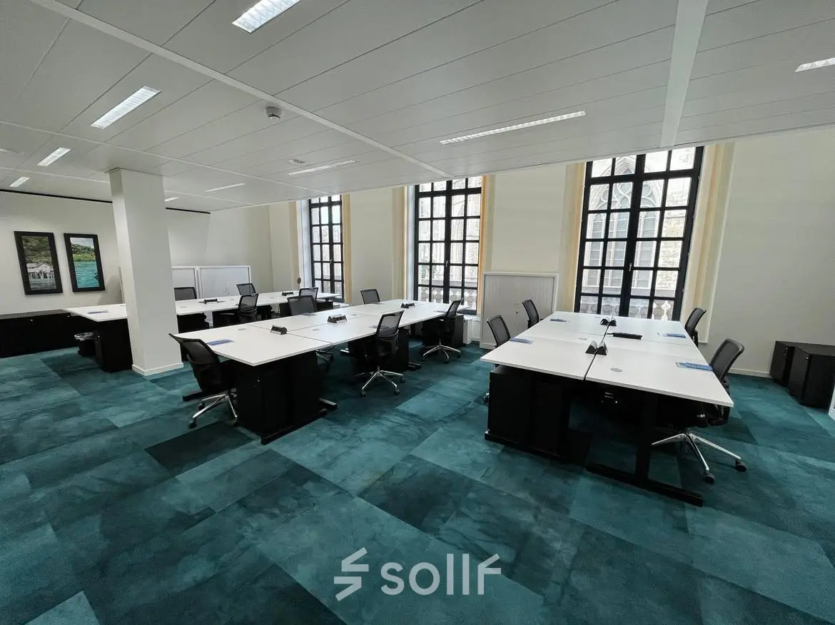 Ruime kantoorruimte te huur op Rue des Colonies 56, Brussel Europese Wijk, met grote ramen en blauwe vloertapijten. Verschillende bureaus en stoelen klaar voor gebruik in een lichte omgeving.
