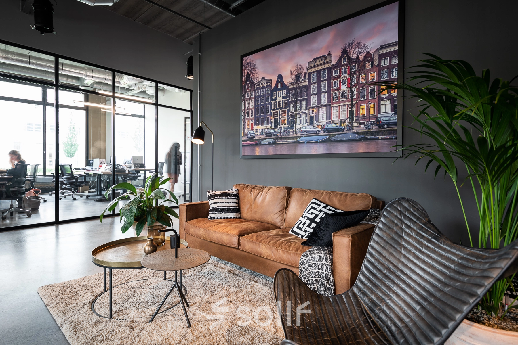 Kantoor te huur in Leuven met moderne, gemeubileerde zithoek, inclusief een bruine leren bank, bijzettafels en groene planten. Op de achtergrond zijn mensen aan het werk in een glazen ruimte.