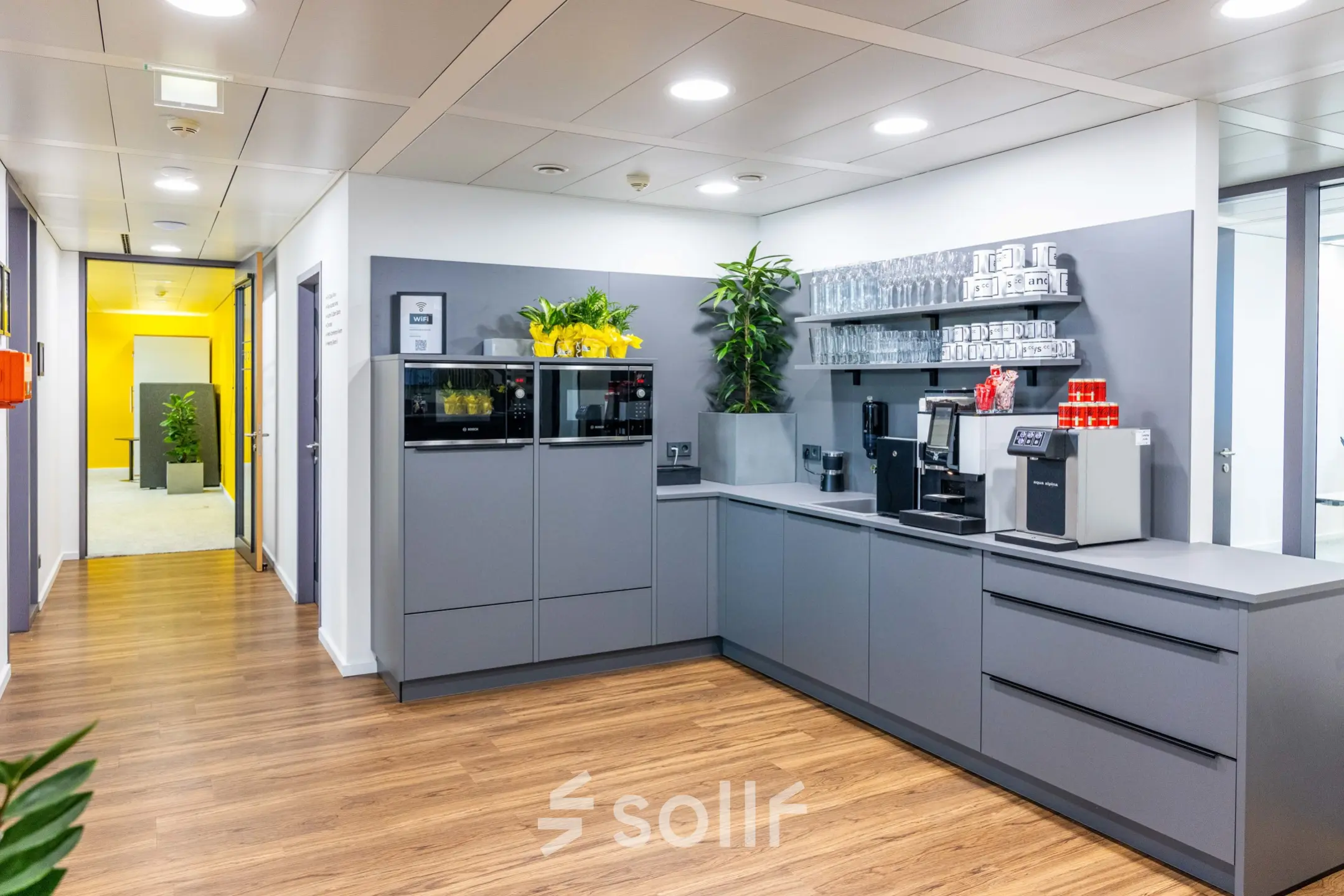 Modern office space featuring a well-equipped pantry with coffee machines and shelving, complemented by indoor plants and sleek wooden flooring at Schönbrunner Strasse 230, 1120 Vienna, ideal for office space rental.