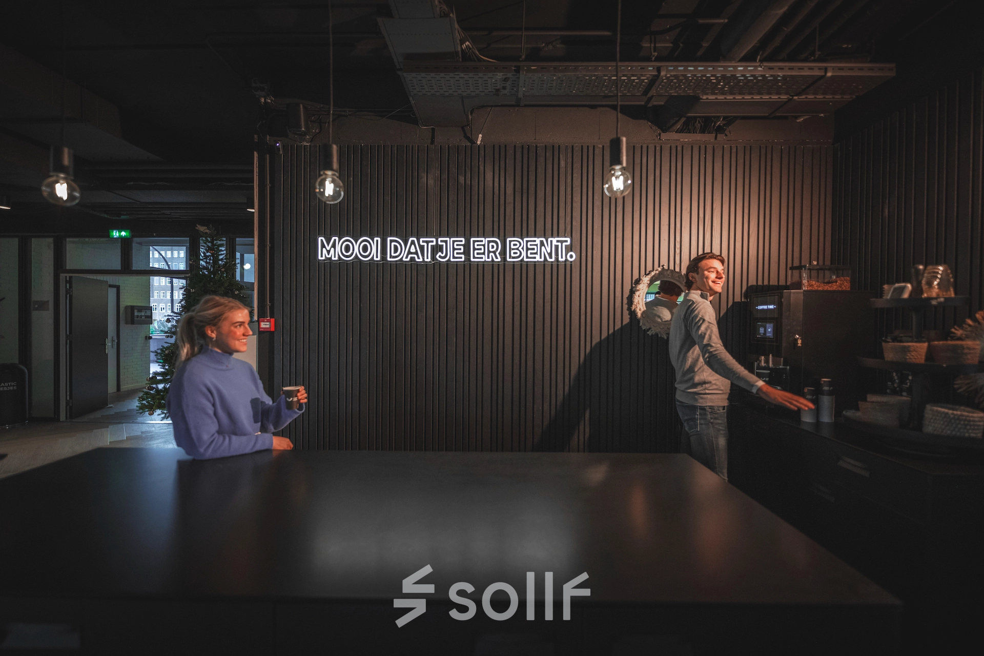 Two people in a modern office space at Egbert Gorterstraat 2-14, Almelo, enjoying coffee near a sleek counter with ambient lighting, ideal for informal meetings. Office space rental available in this vibrant location.