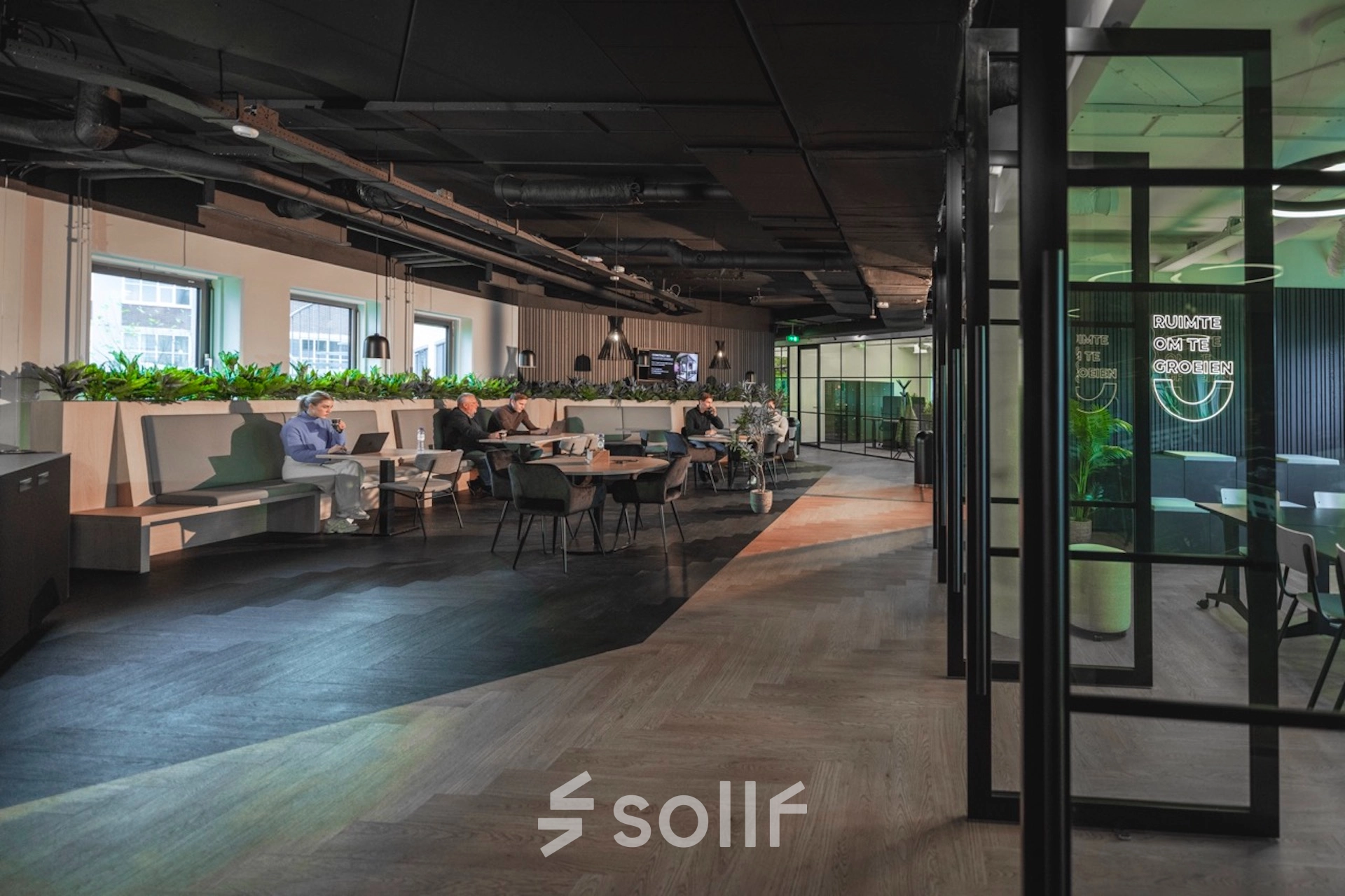 Spacious office interior at Egbert Gorterstraat 2-14 in Almelo, featuring modern workspaces with seating areas, plants, and individuals engaged in business activities. Ideal office space rental for dynamic professionals.