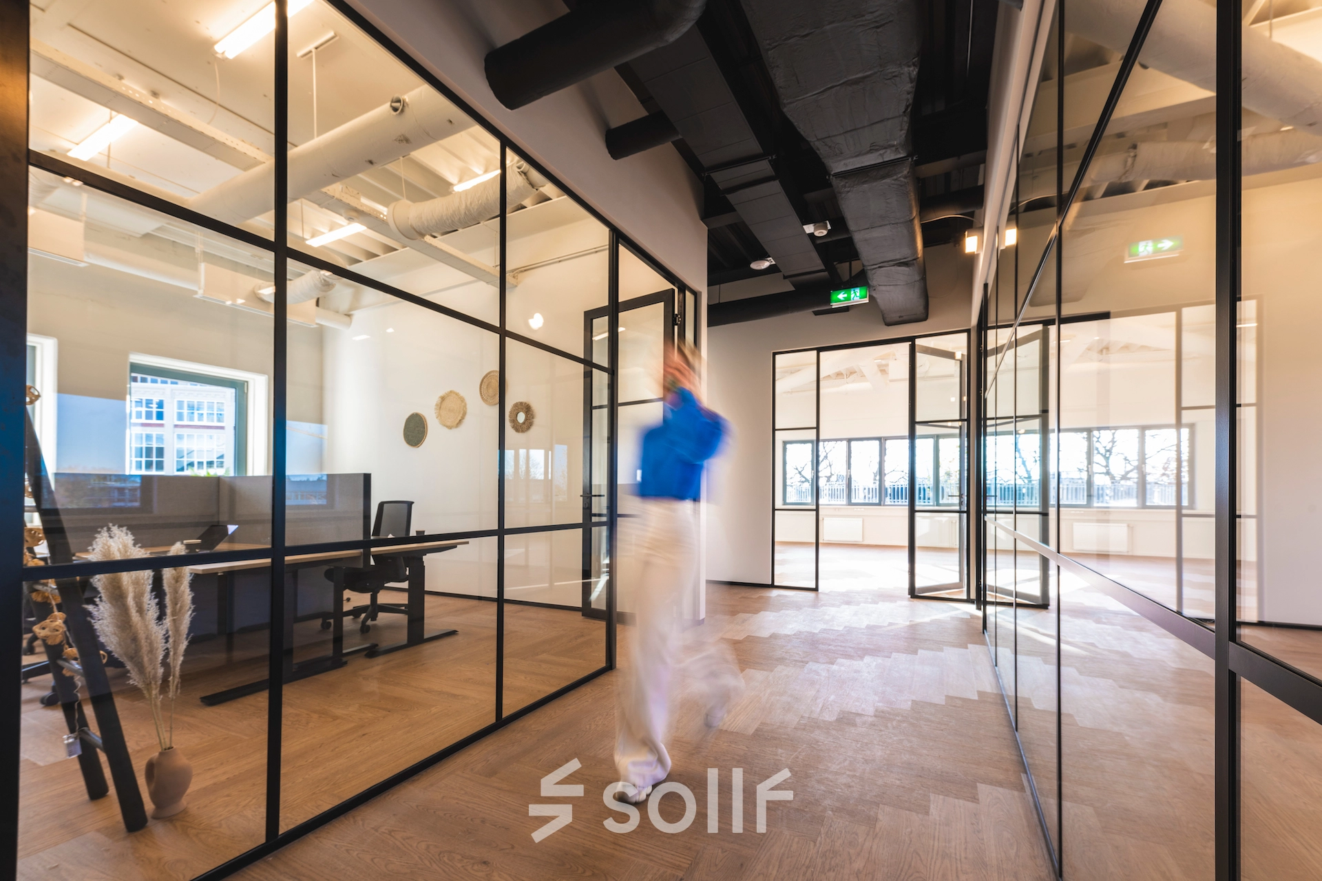 Modern office interior featuring glass partitions and wooden flooring in Egbert Gorterstraat 2-14, Almelo. Sleek design enhances work environments for office space rental, with visible workstations and natural lighting.