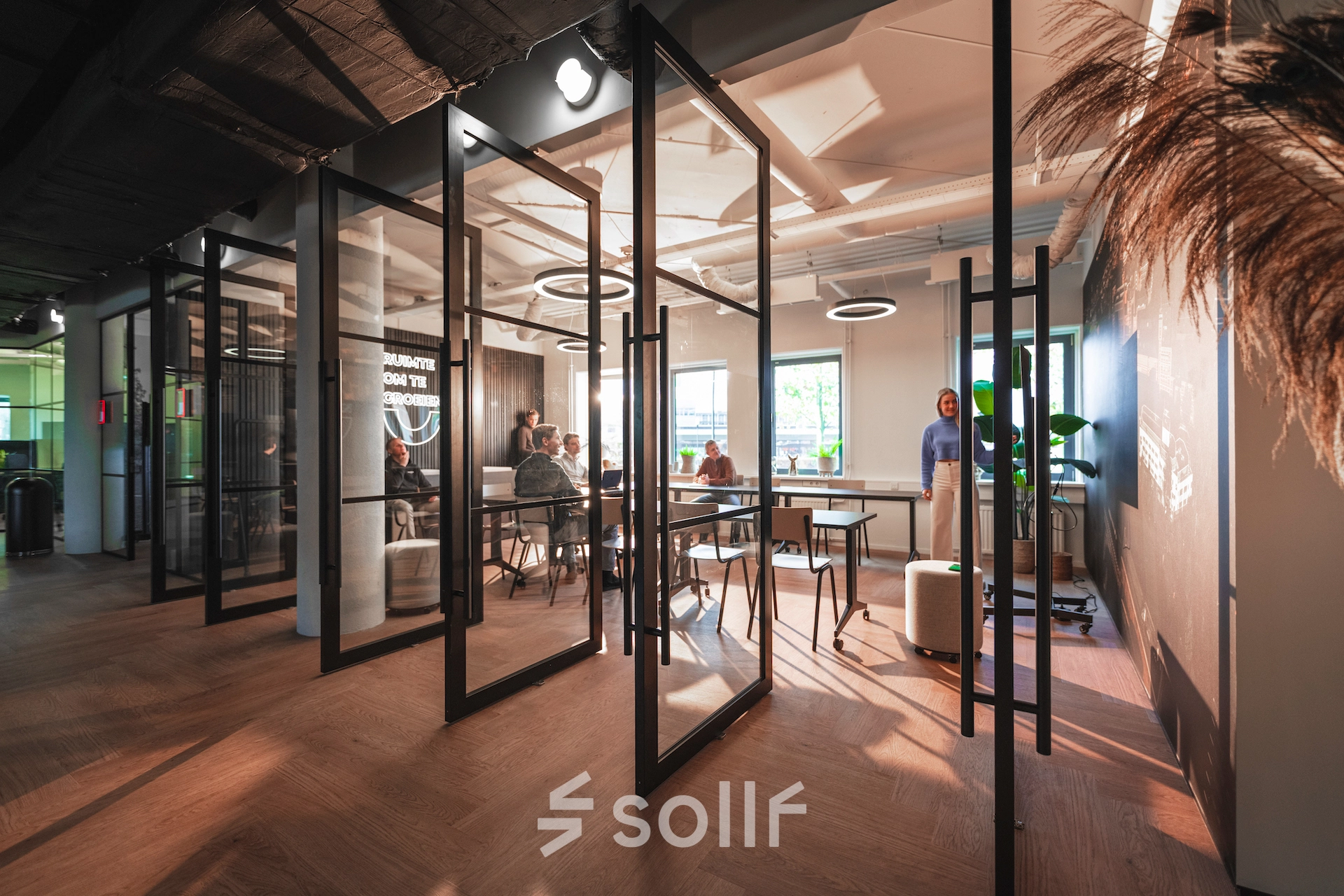 Modern office space rental at Egbert Gorterstraat 2-14, Almelo, featuring glass-walled work areas with people engaged in business discussions under stylish lighting.