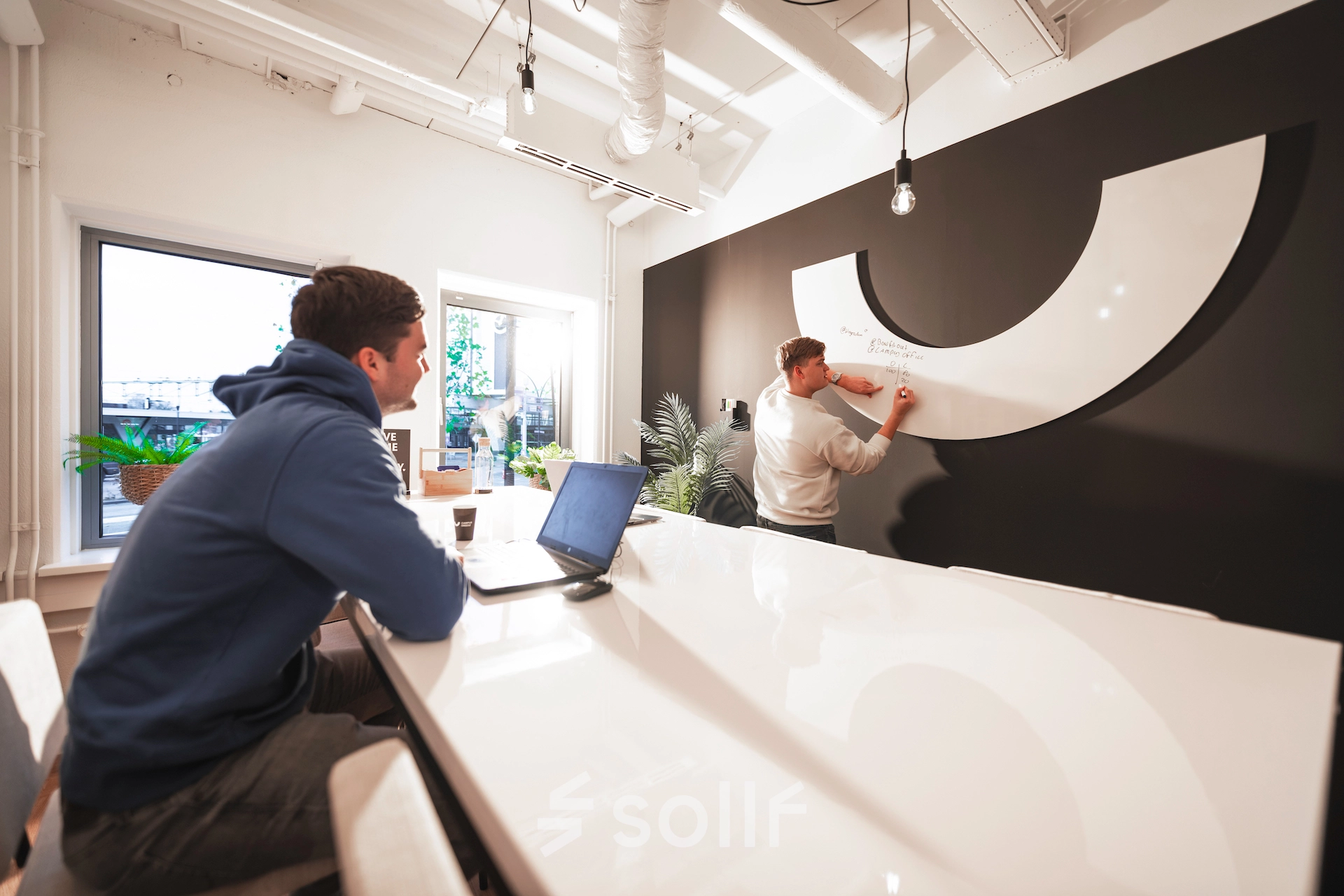 Modern office space rental in Almelo with a sleek interior, featuring a collaborative work environment. Two professionals engage in brainstorming at a high gloss table with a stylish black and white backdrop.