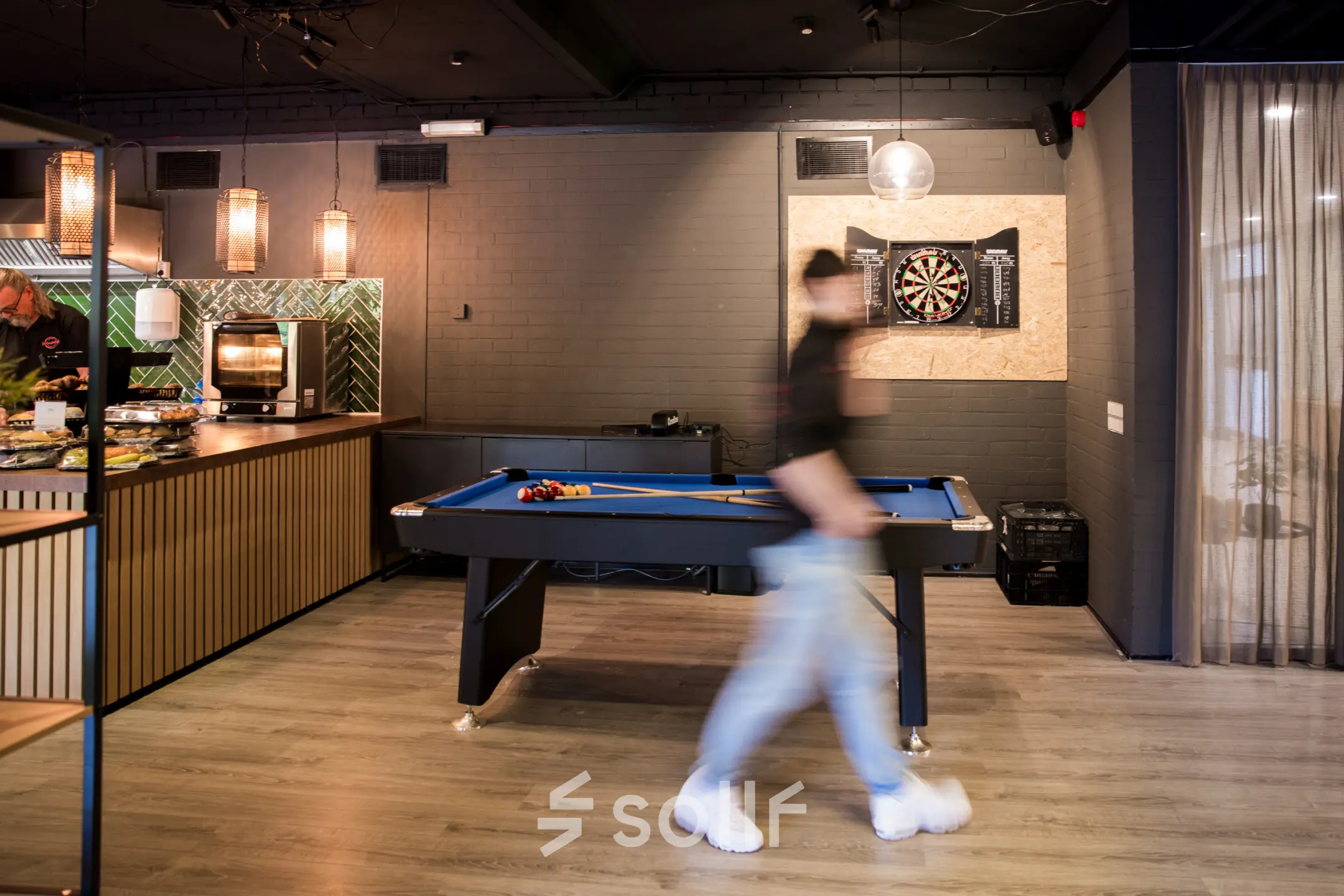 Modern office lounge area with a pool table and a person walking by, featuring stylish lighting and a small kitchen setup in Almere Stad office space rental.
