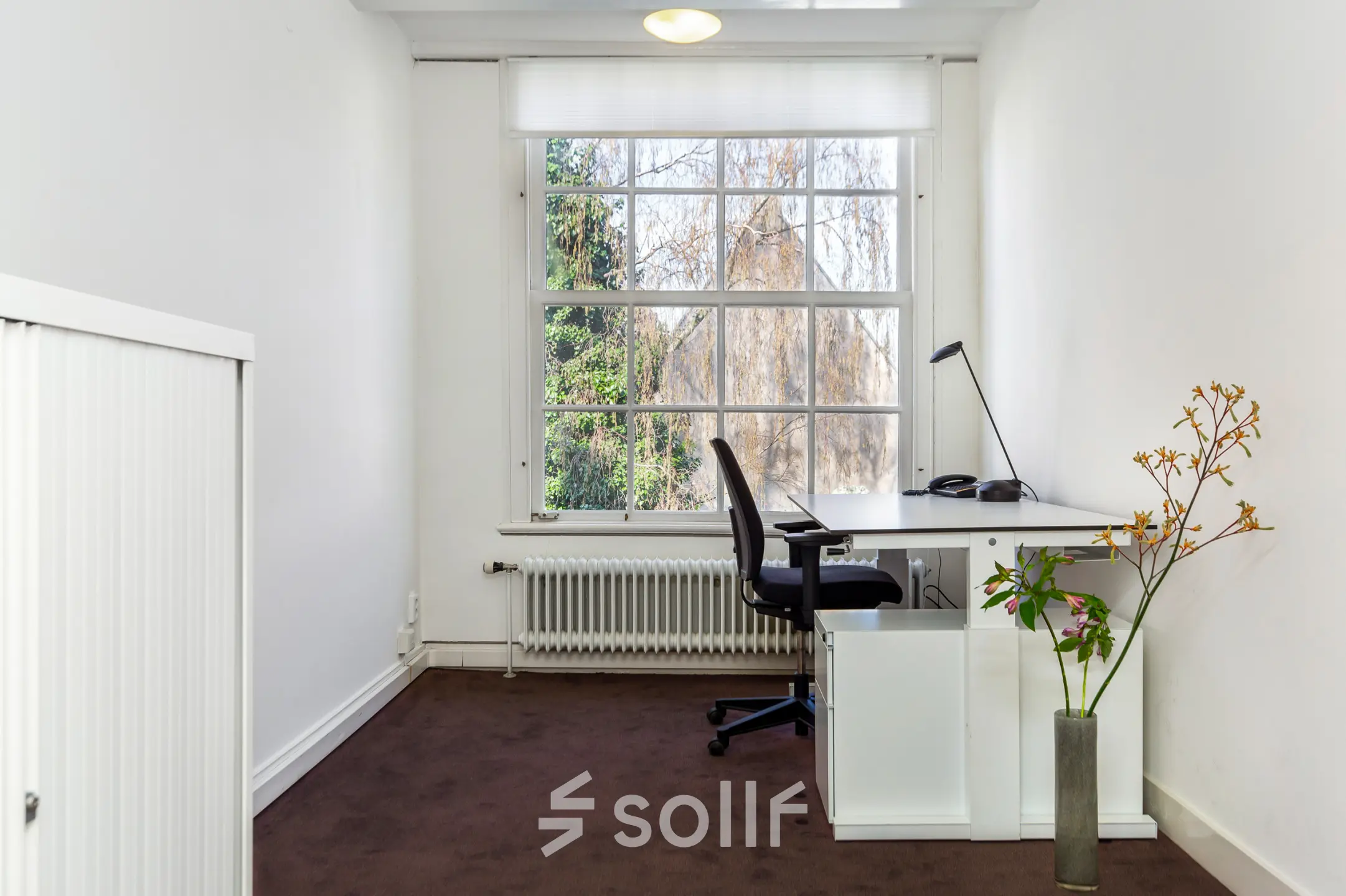 Bright furnished office space for rent on Keizersgracht 62–64 in Amsterdam Canal Belt, featuring a desk with a chair and large window overlooking trees.