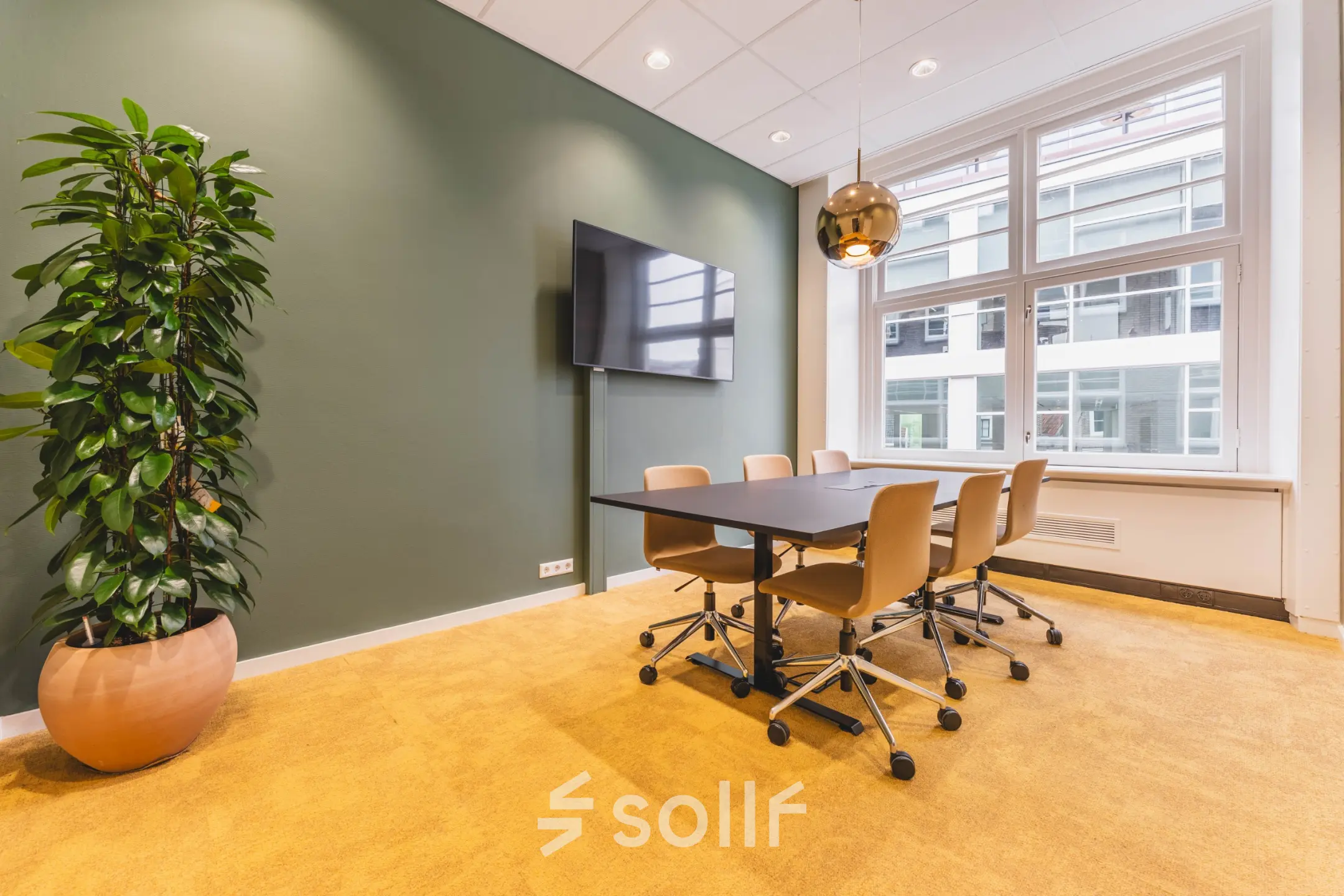 Bright office space for rent at Herengracht 471, Amsterdam Center, featuring a meeting table, chairs, wall-mounted TV, and large window with natural light.