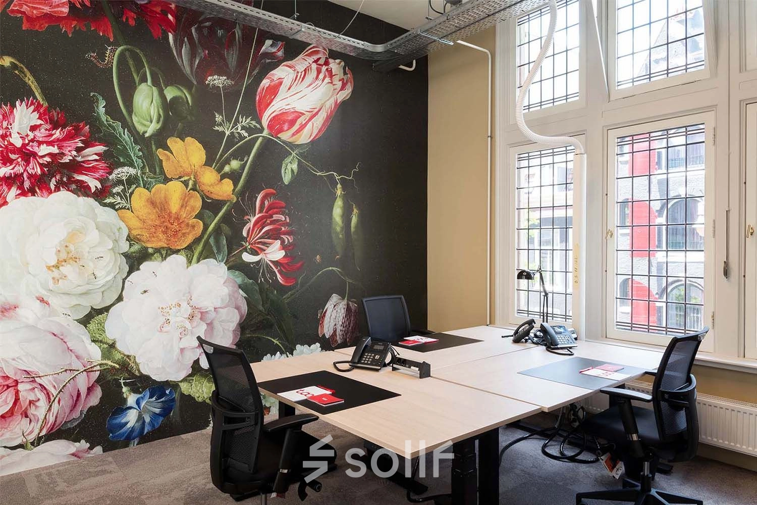 Bright office space at Nieuwezijds Voorburgwal 104-108, Amsterdam Center, featuring a bold floral mural and well-lit workstations for rent.