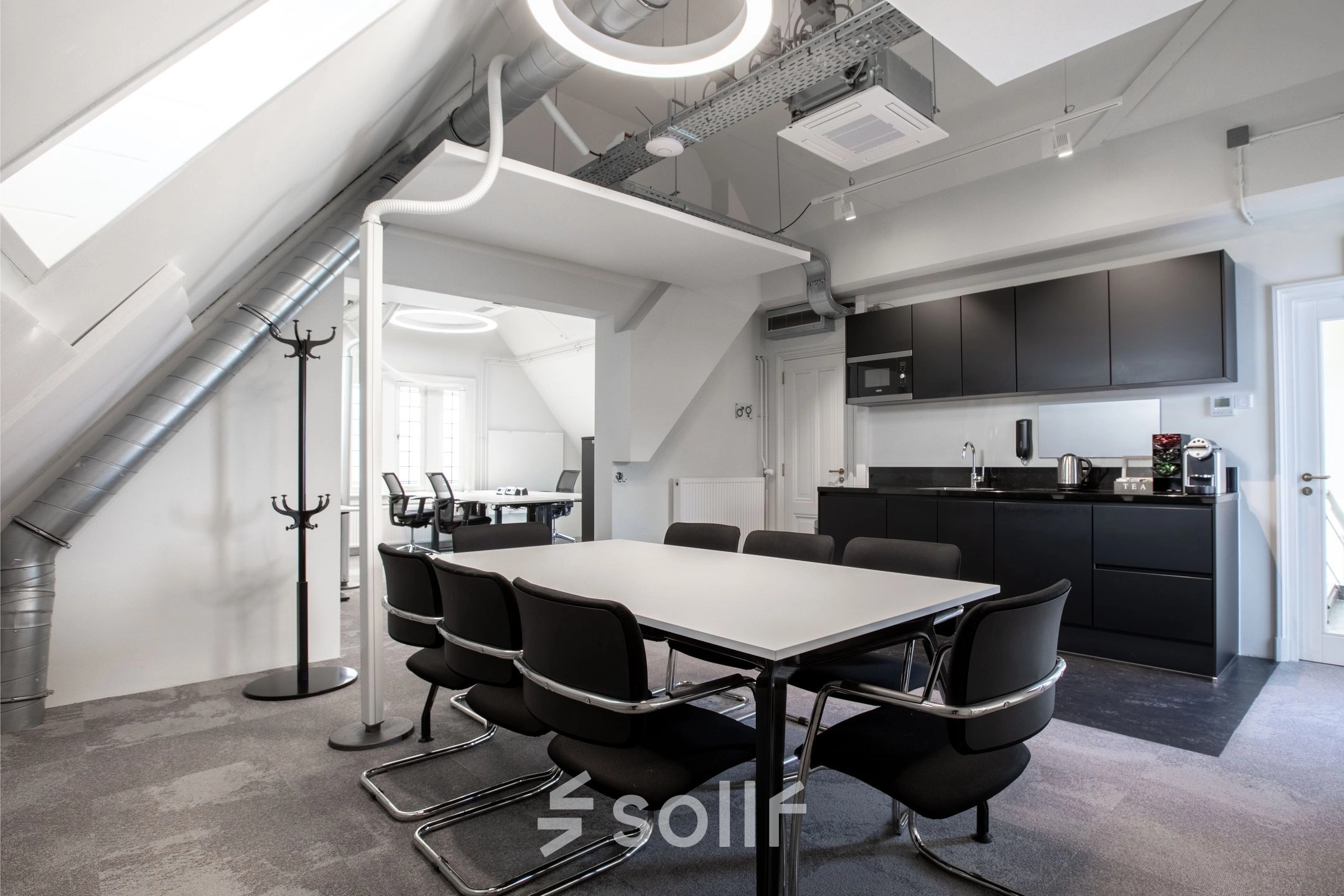 Modern office to rent at Nieuwezijds Voorburgwal 104-108, Amsterdam Center, featuring a sleek meeting table, black chairs, and a kitchenette. Ideal for collaborative work and meetings.