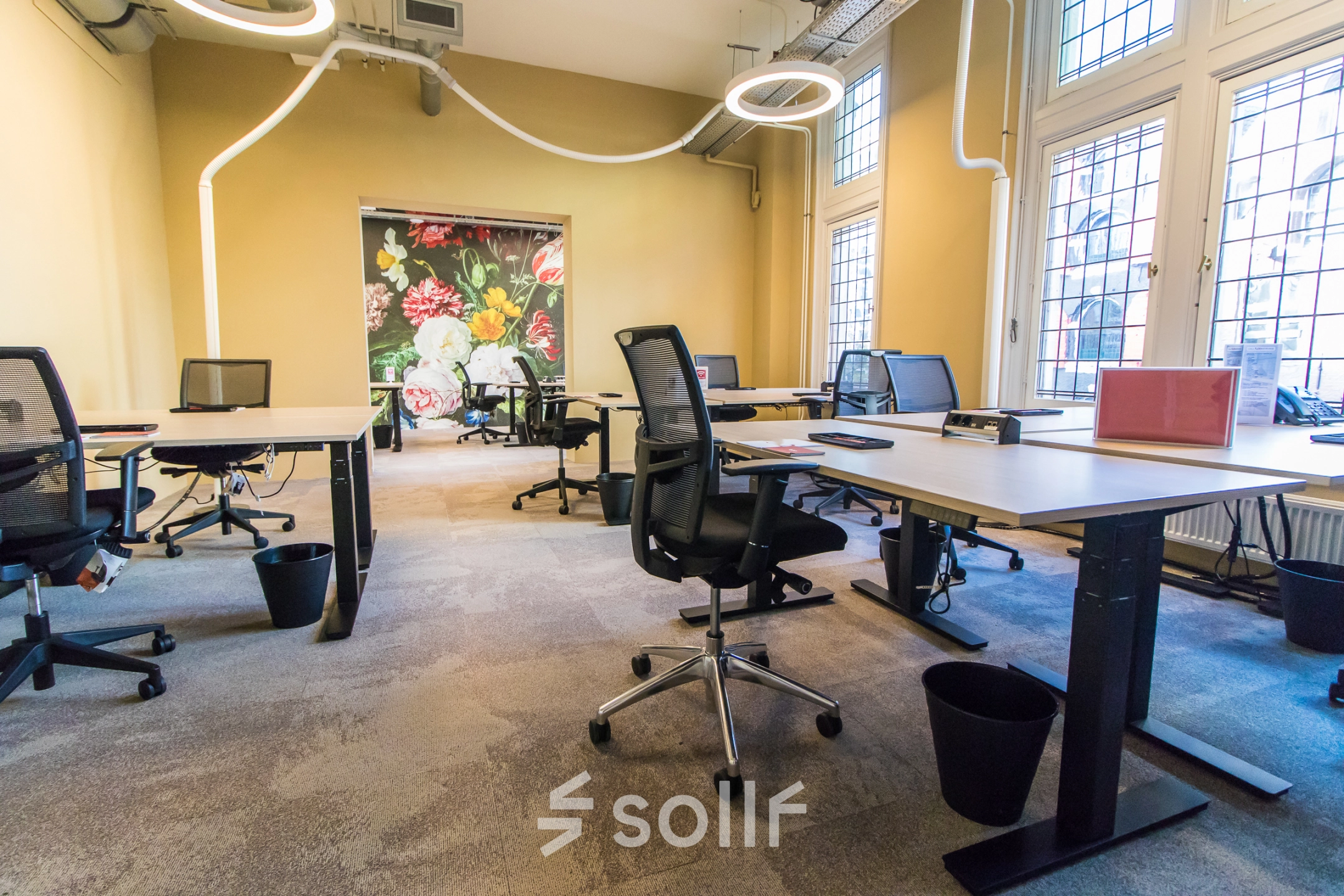 Bright office space with modern workstations and charming floral wall art, available for rent at Nieuwezijds Voorburgwal 104-108, Amsterdam Center. Perfect for dynamic and creative professionals seeking office space rental.