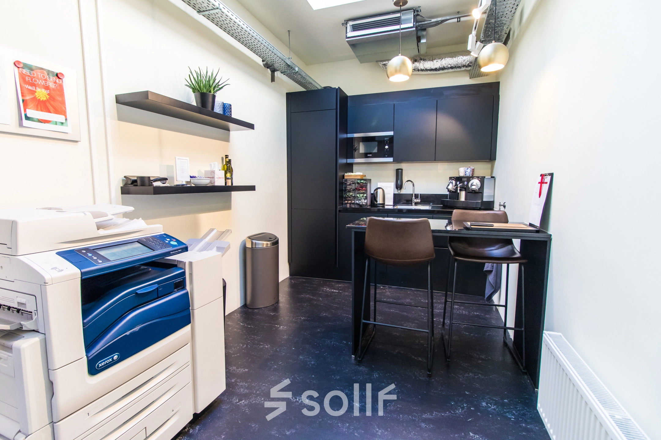 Compact, furnished kitchenette in an Amsterdam office, featuring a high table with chairs, appliances, and a printer. Ideal for small office space rental.