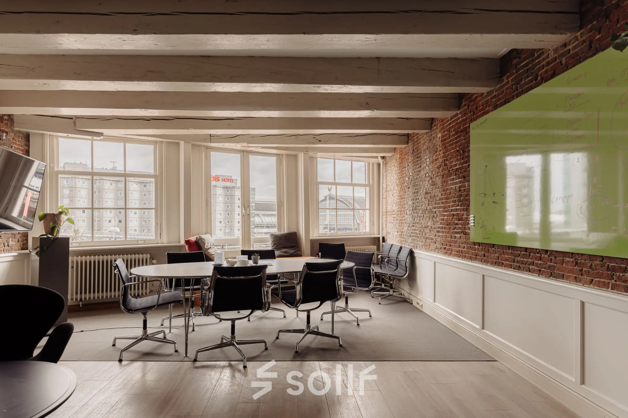 Charming office space at Prins Hendrikkade 14 in Amsterdam Center with exposed brick walls, a large table, modern chairs, and a large window with city views. Ideal for office space rental.