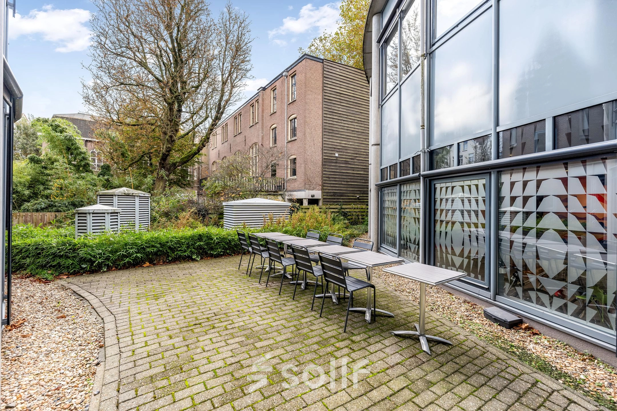 Outdoor view of office space rental at Stadhouderskade 55 in Amsterdam Center featuring a brick patio with tables and chairs, surrounded by greenery.