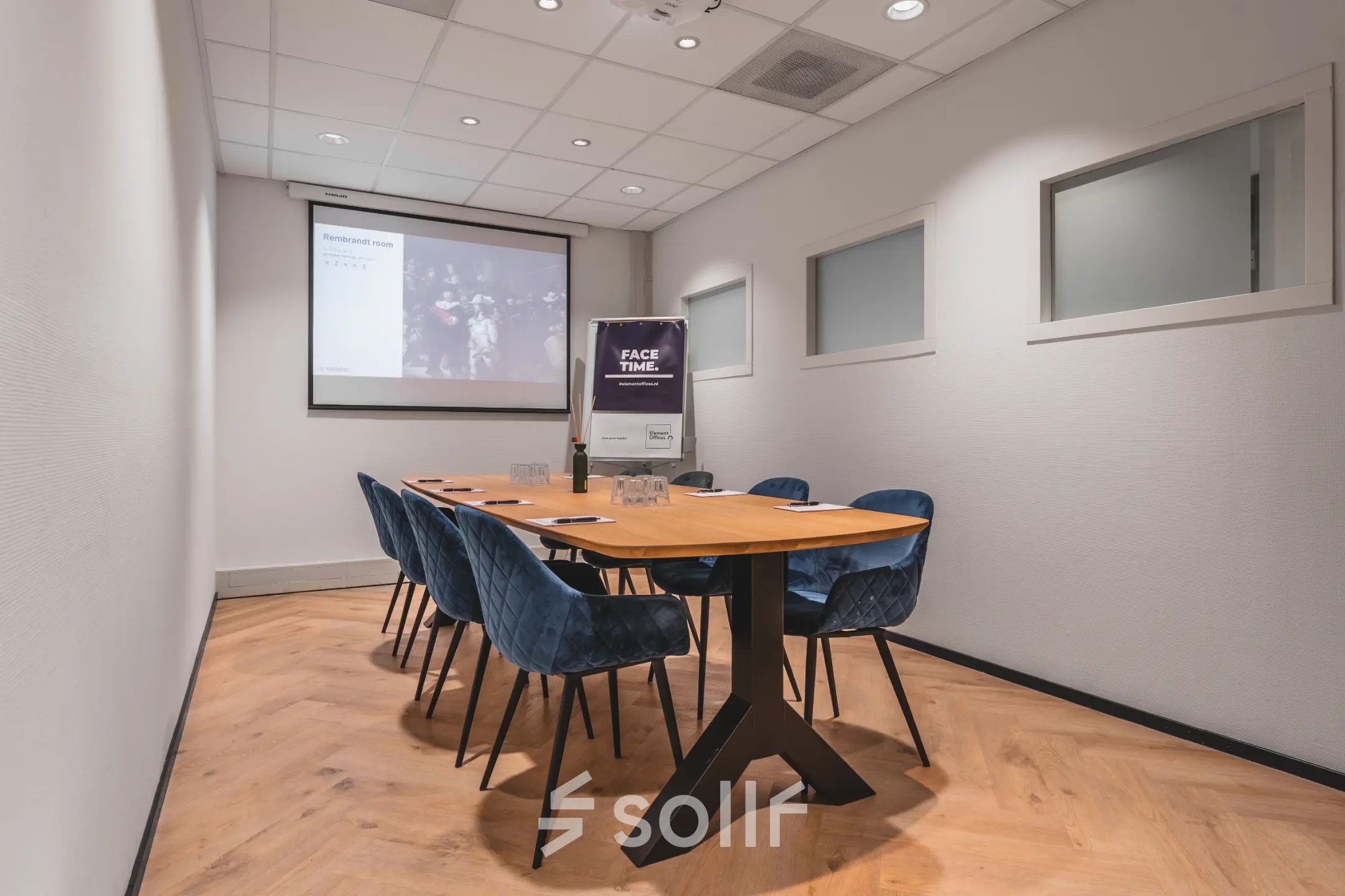A conference room featuring a sleek wooden table with blue chairs, a large screen, and projector setup, perfect for office space rental in Amsterdam Duivendrecht.