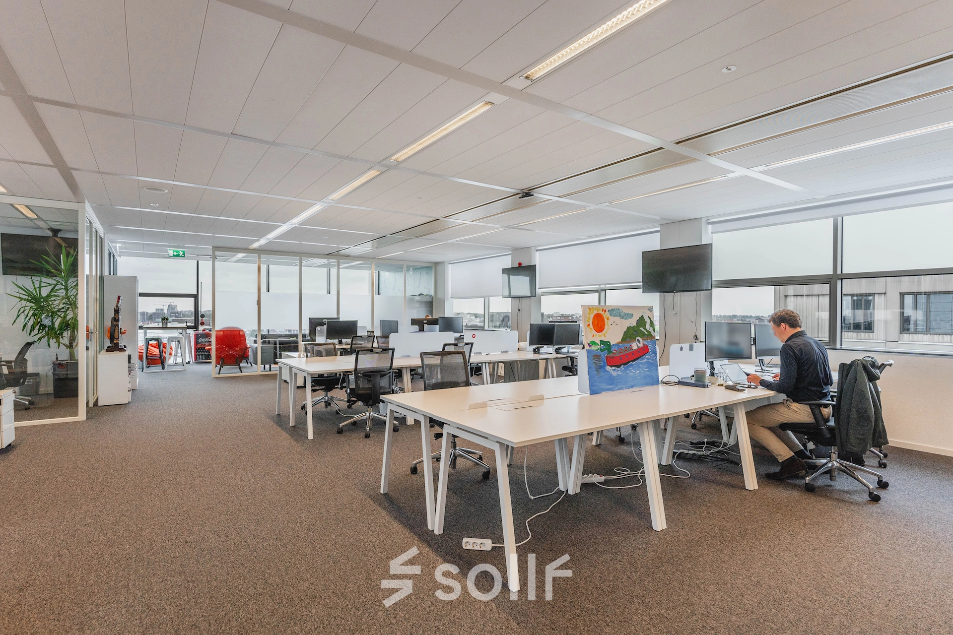 Spacious and well-lit office space rental at Piet Heinkade 95B in Amsterdam East, featuring sleek workstations and modern furnishings. A person is working at a desk by the window.