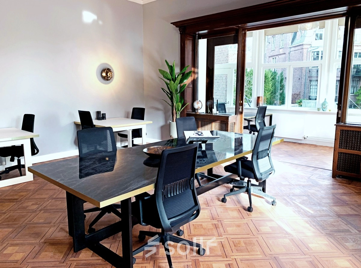 Bright office space in Amsterdam Old South at Emmalaan 7-9, featuring modern desks and chairs available for rent. Large windows allow natural light, ideal for productive work environments.