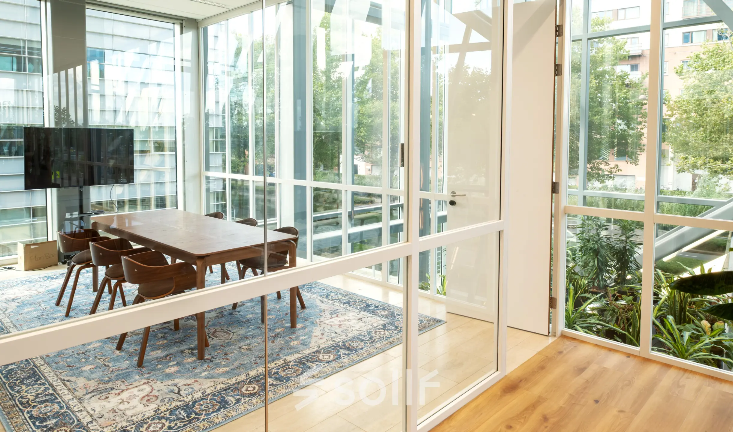 Bright office space with a large wooden table and chairs surrounded by glass walls offering views of greenery, ideal for office space rental.