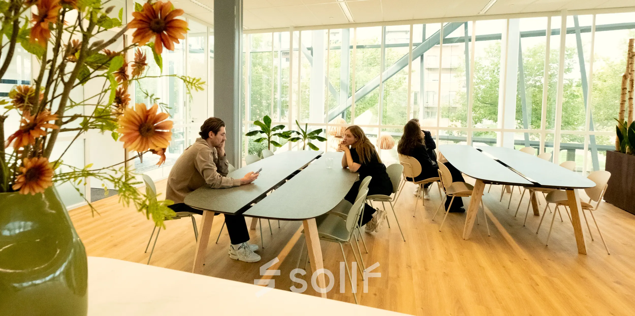 Office space rental in Amsterdam Riekerpolder, featuring spacious tables and large windows. People are engaged in conversation, adding a collaborative vibe.