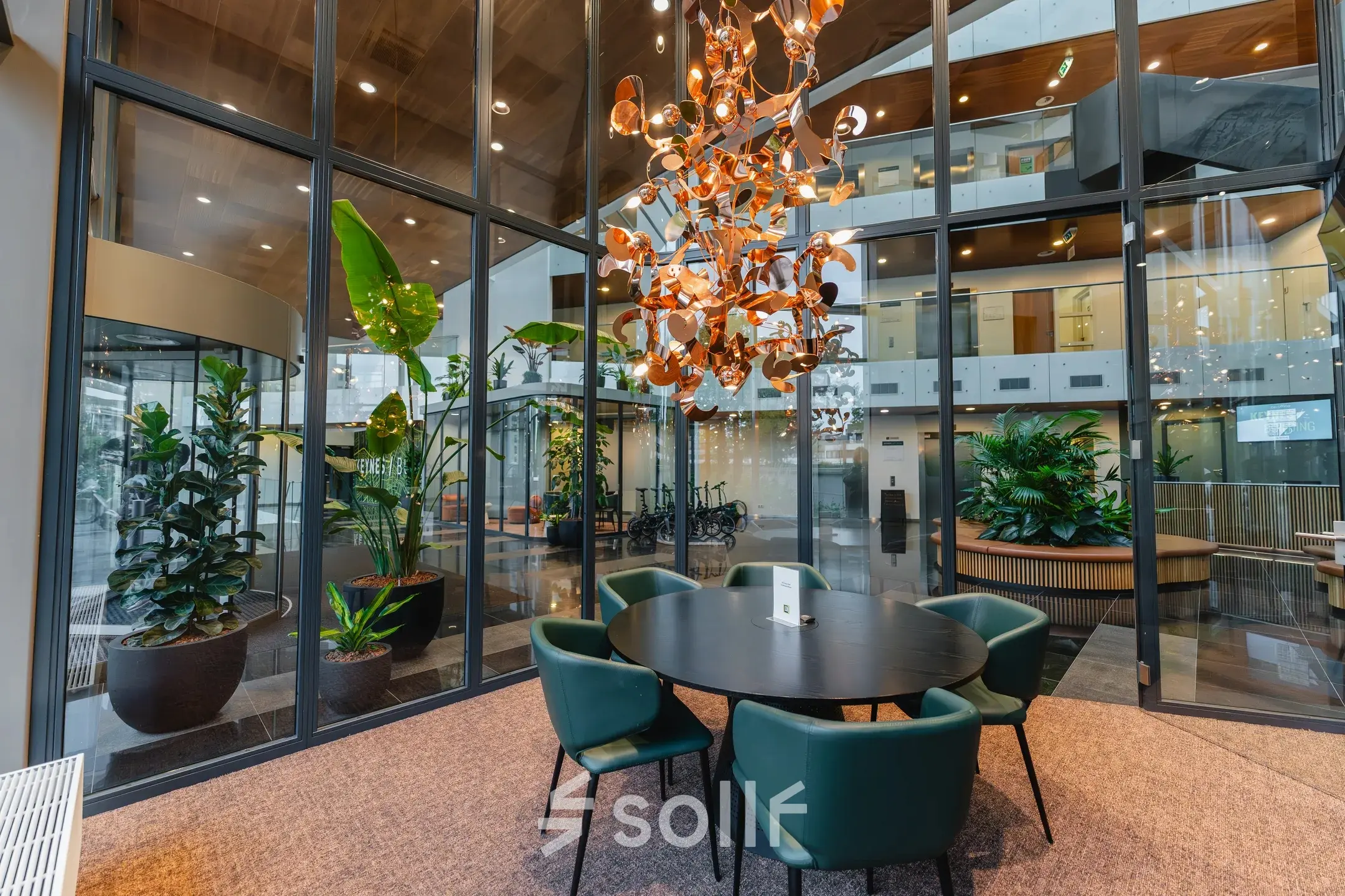 Modern furnished workspace at John M. Keynesplein 4-10, Amsterdam Riekerpolder, with a round table and green chairs beneath an artistic chandelier, surrounded by glass walls and indoor plants.