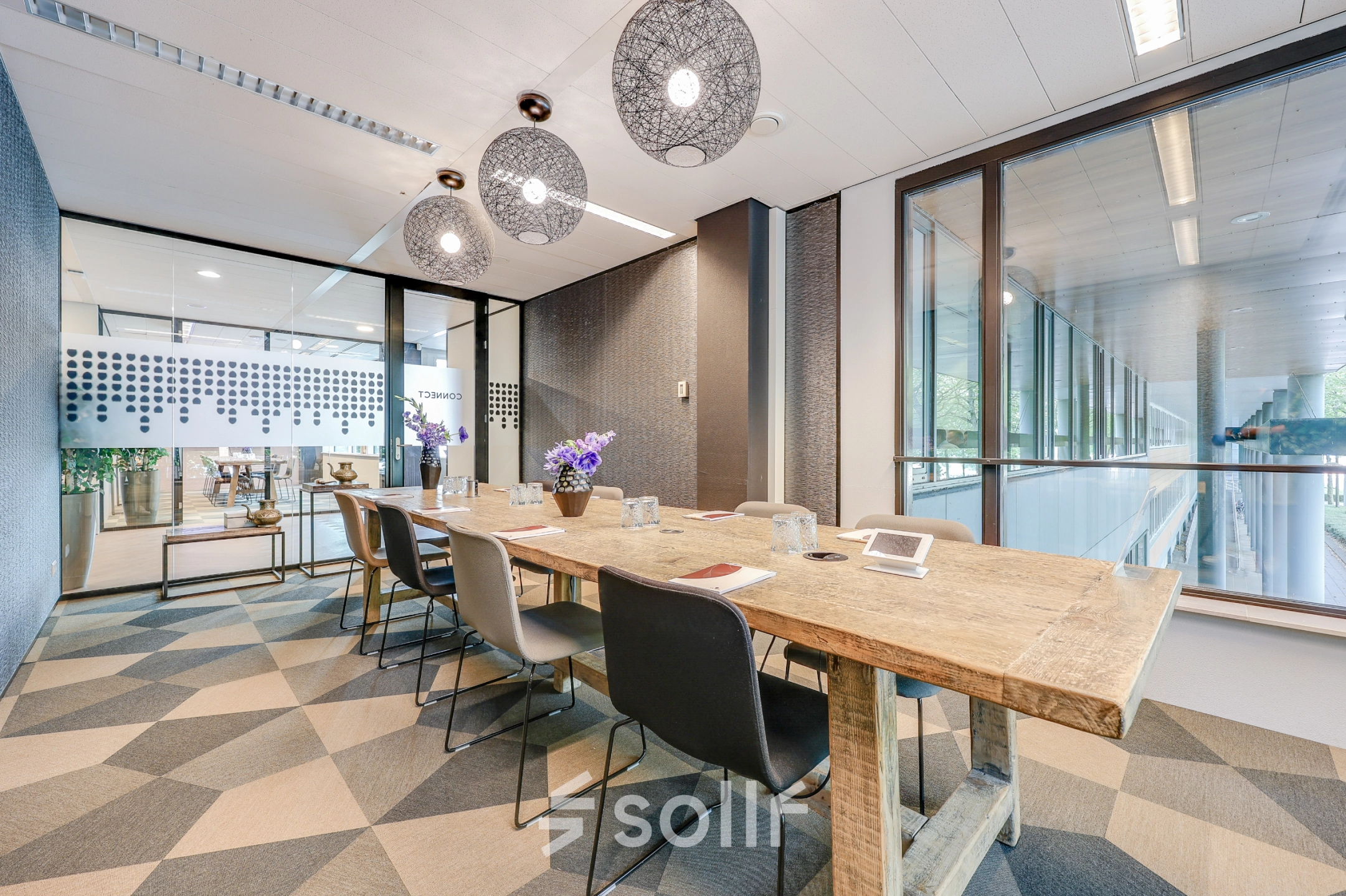 Bright office space in Amsterdam Riekerpolder with a wooden table, modern chairs, and glass partitions, ideal for rent office space. The setting is modern with hanging lights and natural lighting.