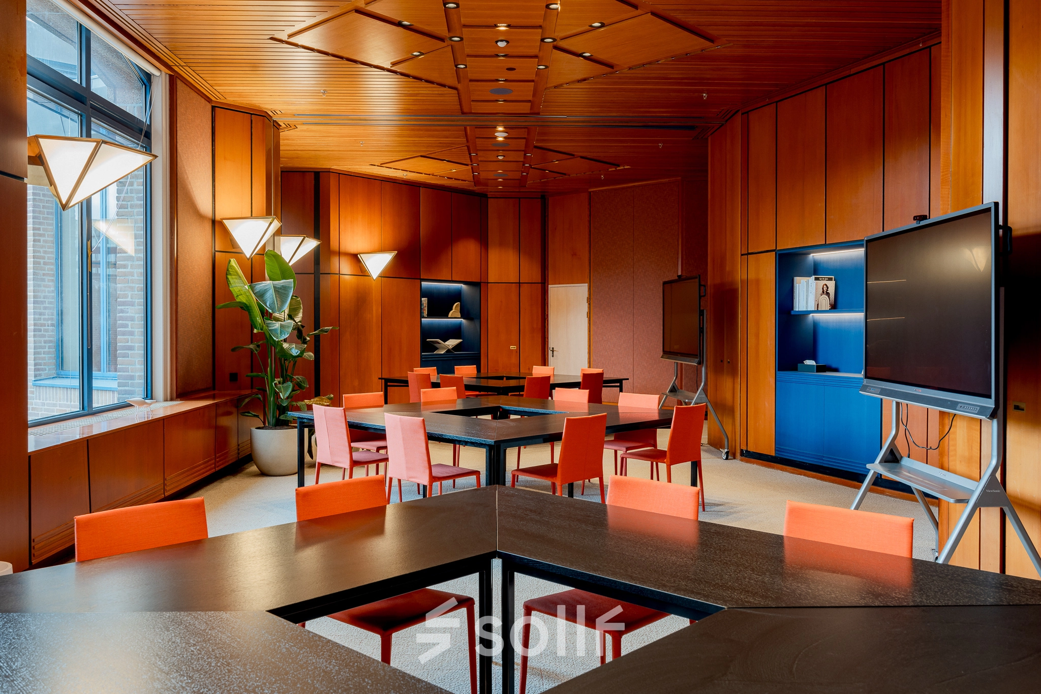 Modern office space with wood paneling, vibrant orange chairs around tables, and large screens, ideal for office space rental in Amsterdam South East.