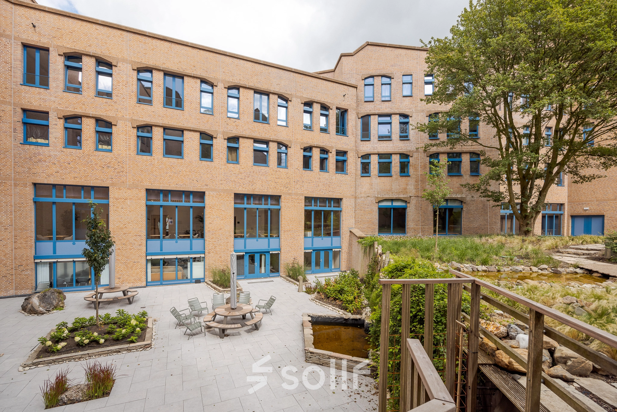 Exterior view of Bijlmerplein 888 in Amsterdam South East, showcasing a brick building and a peaceful patio. Ideal office space rental with outdoor seating options.