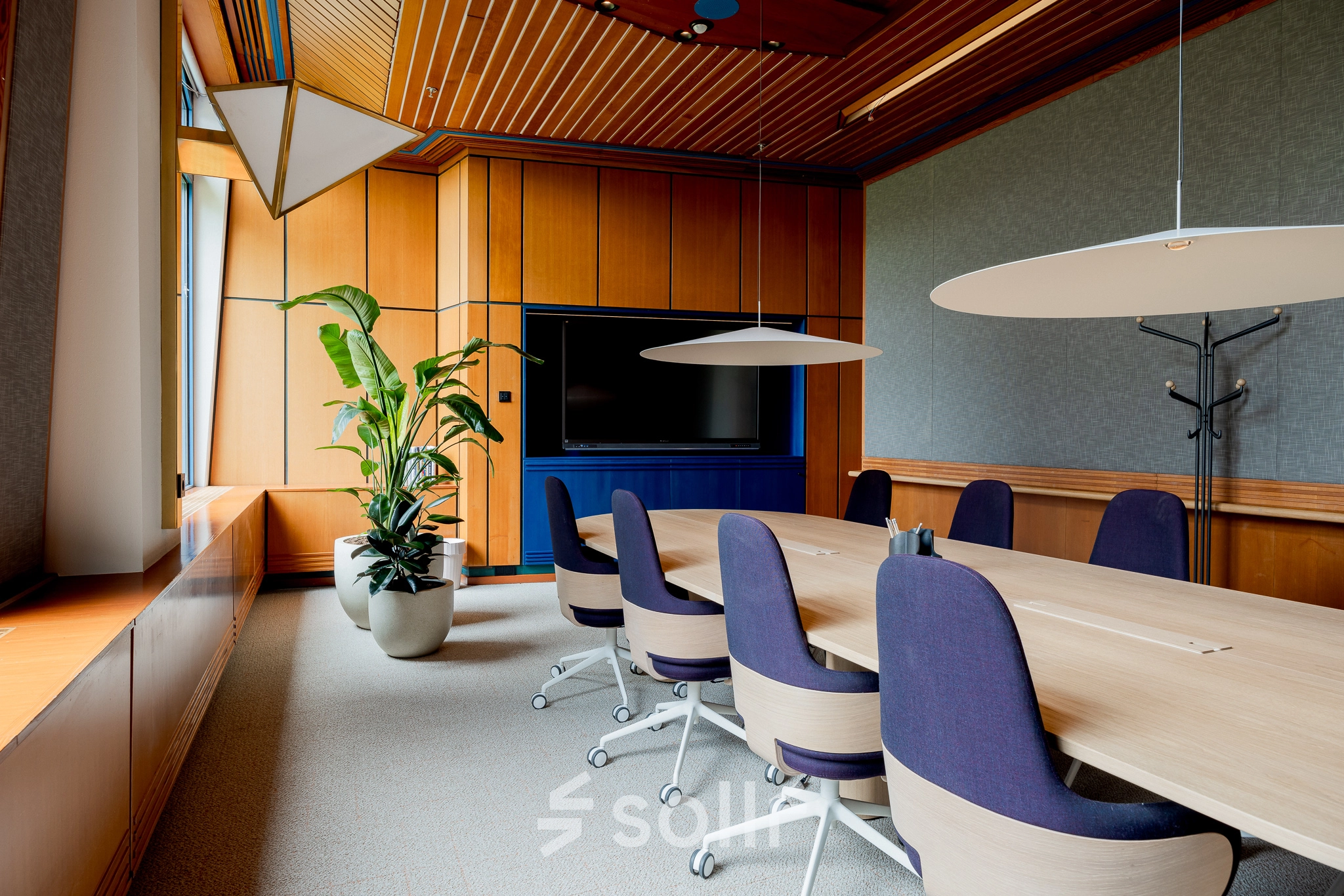 Elegant office space featuring a long wooden table, blue chairs, a large TV, and potted plant at Bijlmerplein 888, ideal for office space rental in Amsterdam South East.