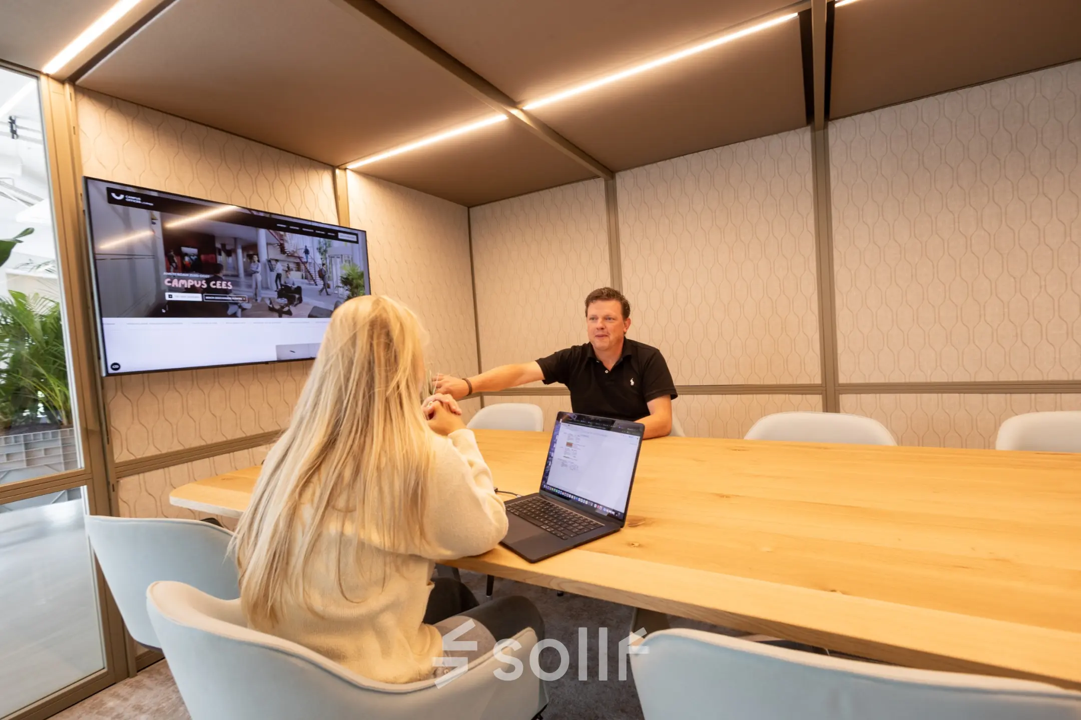 Two people engage in a meeting in a modern, well-lit office space with a large wooden table and digital screen, suitable for office space rental in Amsterdam South East.