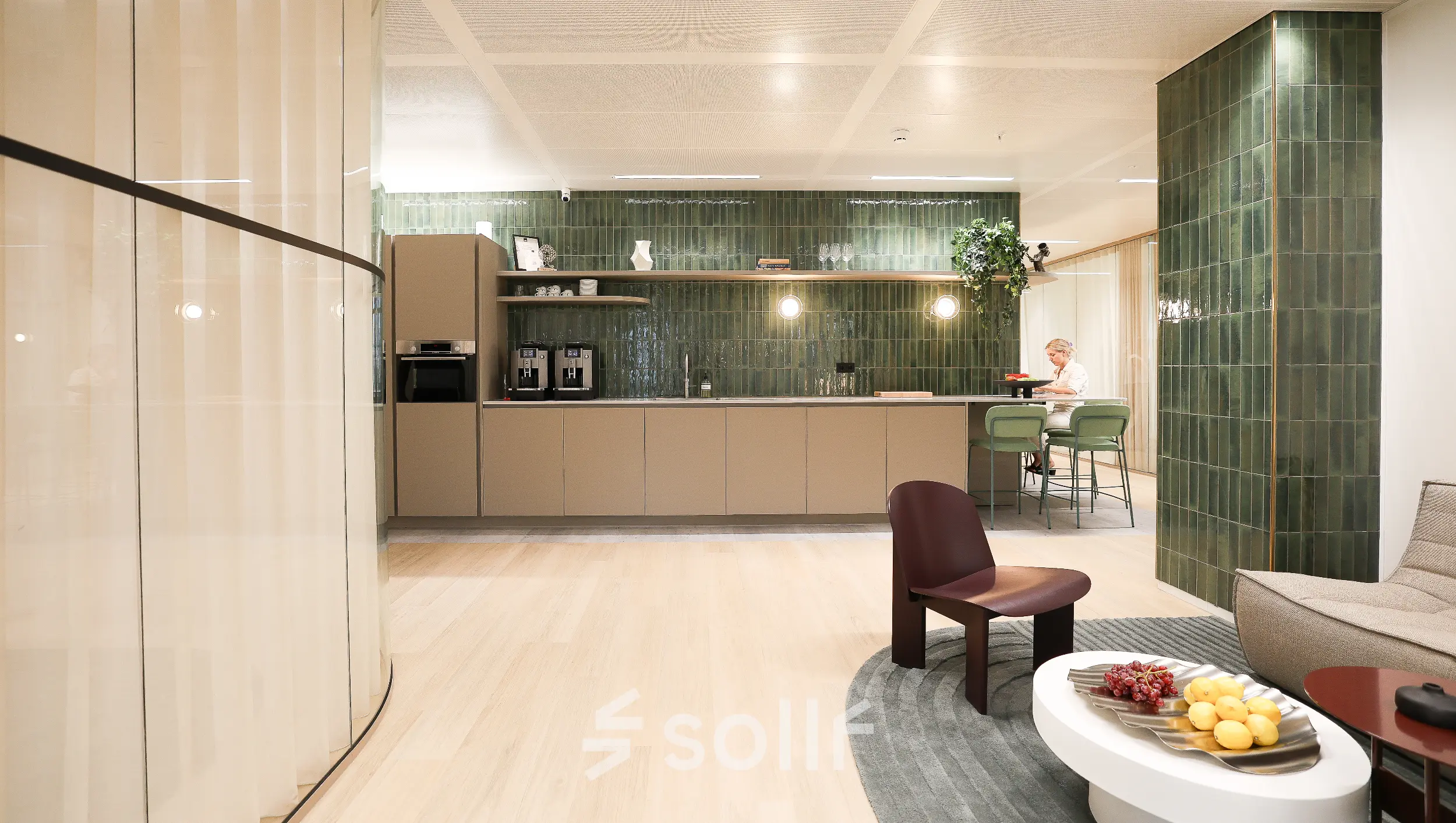 Modern office space available for rent in Amsterdam Zuidas, featuring a furnished lounge area with seating and a kitchenette. A person is engaged in a business activity at a table.