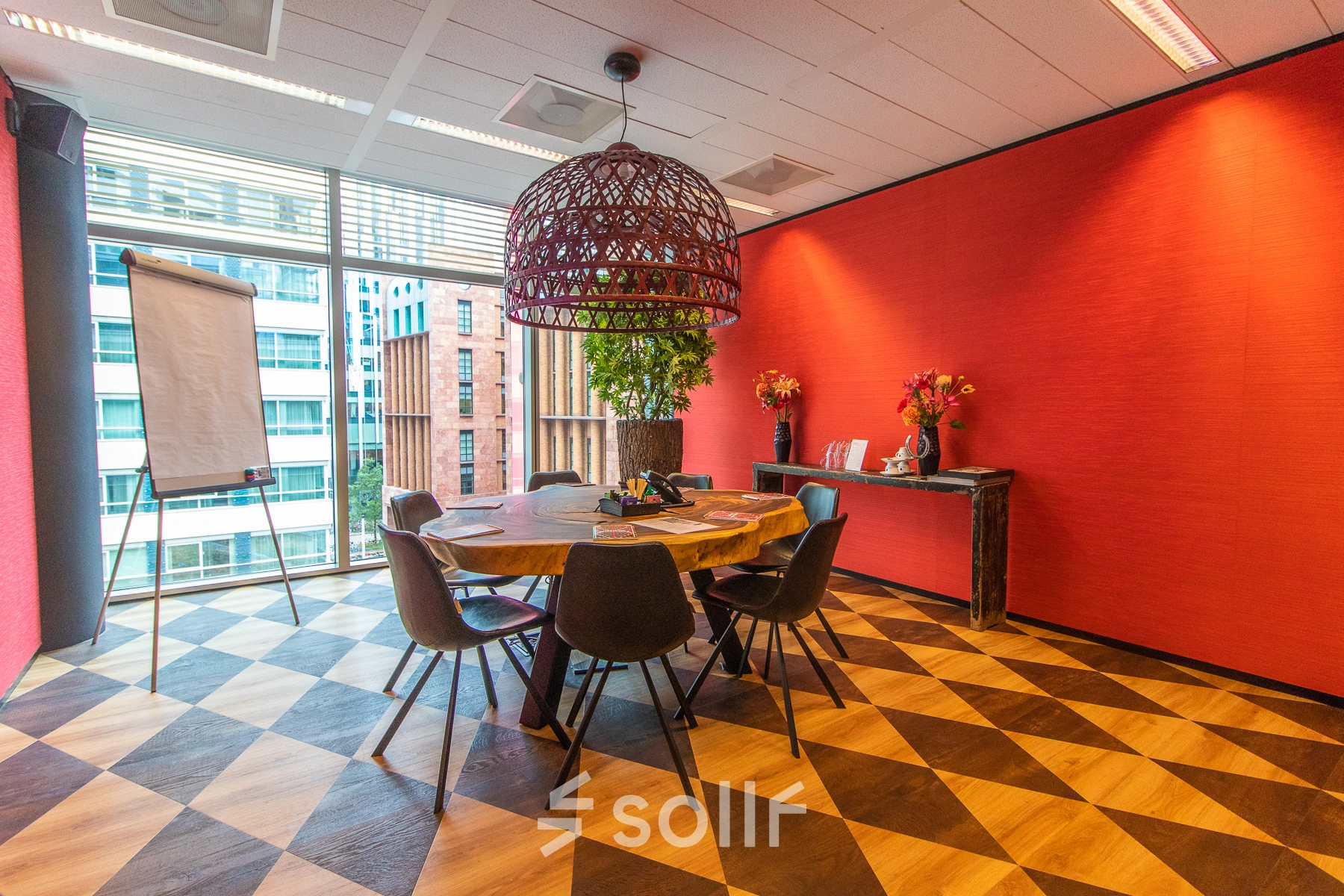 Bright conference room in Amsterdam Zuidas, featuring a large round table with chairs, vibrant red walls, and a large window overlooking a cityscape. Ideal for office space rental.