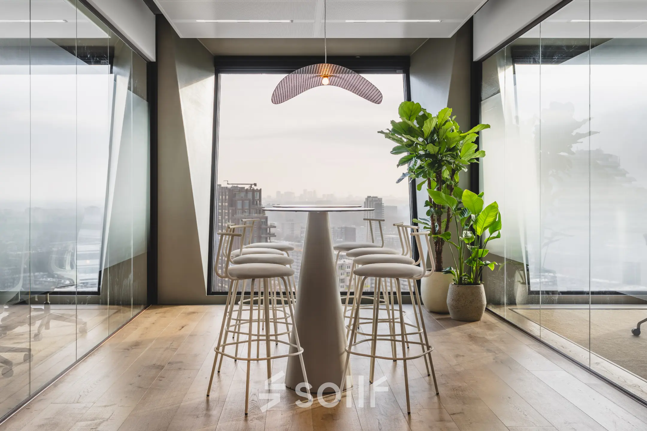 A modern office space with tall windows in Amsterdam Zuidas, featuring high stools around a central table, ideal for collaborative meetings. Rent office space with stunning city views.