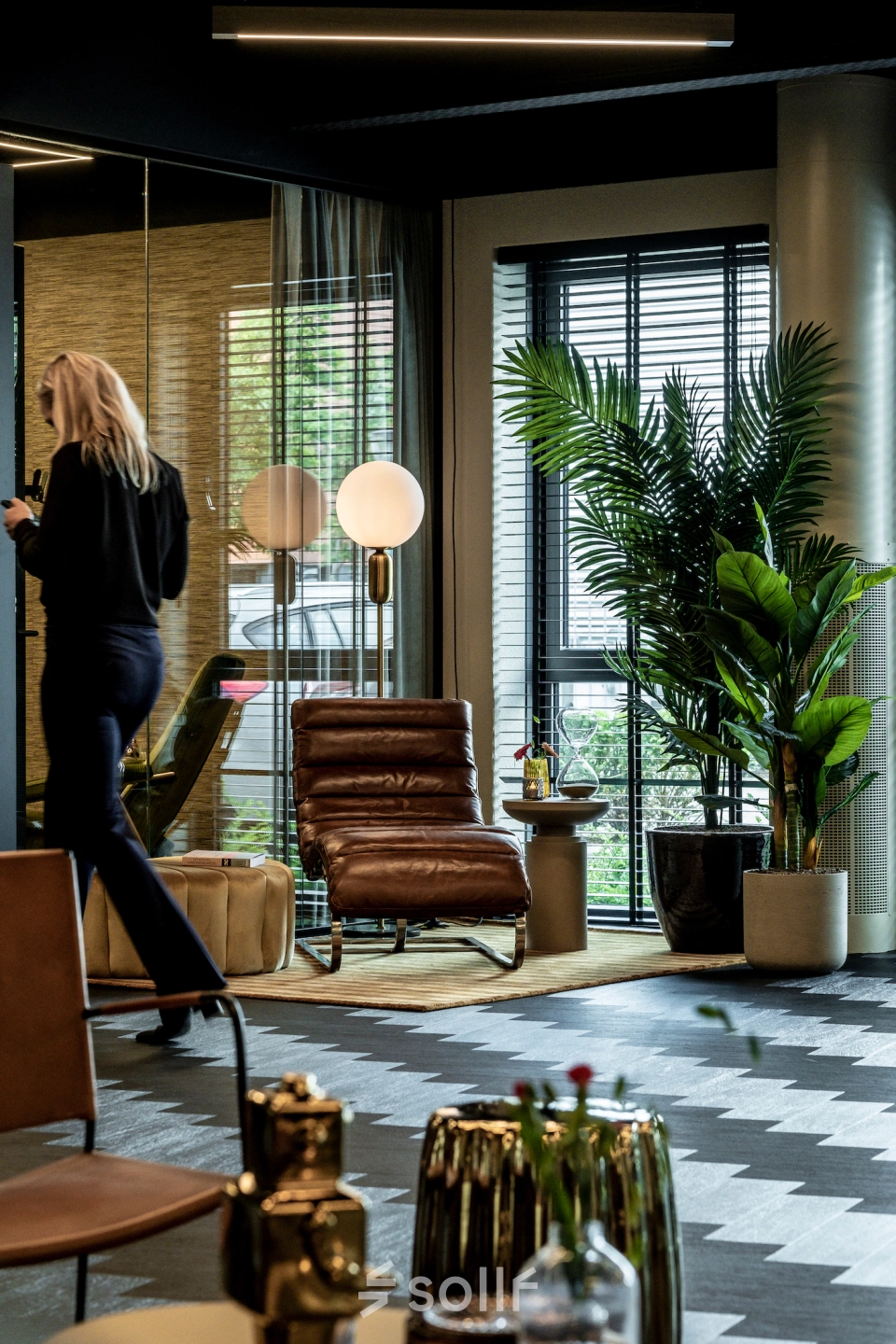 Modern furnished office space in Apeldoorn with stylish seating and plants. A person is walking while using a smartphone, suggesting business activity.