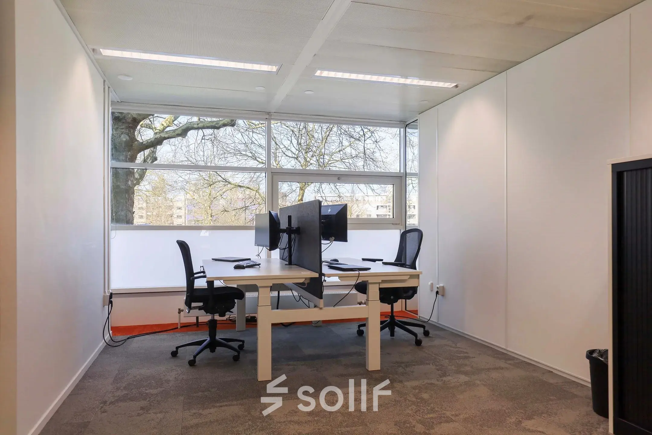 Bright office space for rent in Apeldoorn, featuring two desks with chairs and screens, large windows offering natural light.