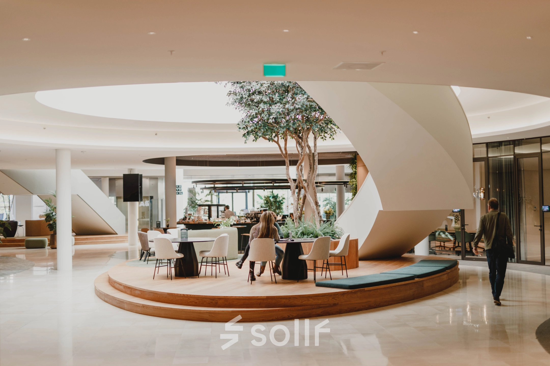 Modern office space to rent at Rijksweg-West 2, Arnhem Zuid, featuring a stylish lounge area with tables and chairs under a skylight. People are working and walking through the space.