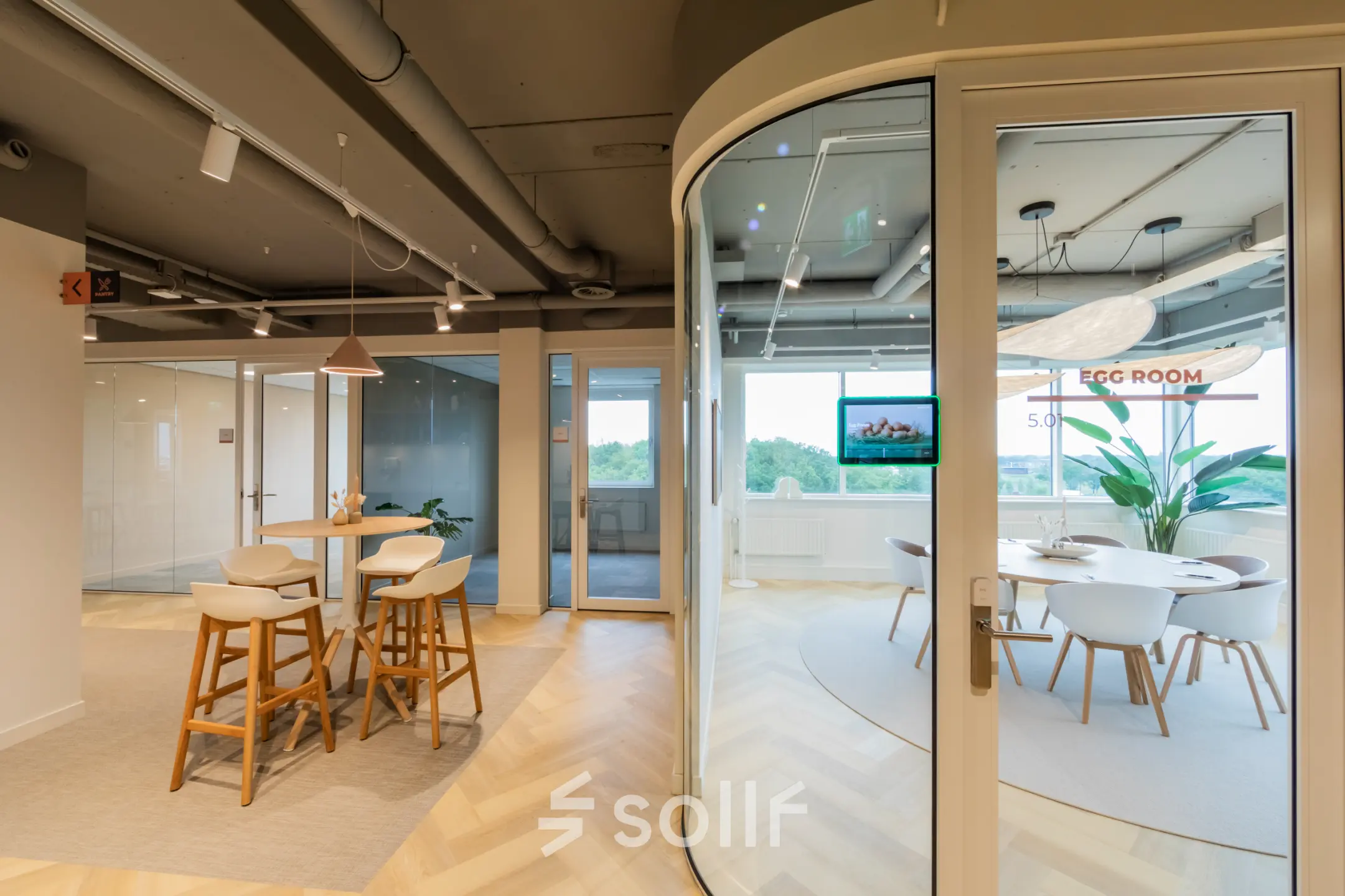 Modern office space for rent featuring glass-walled meeting room, bar stools at a high table, and large windows providing natural light.
