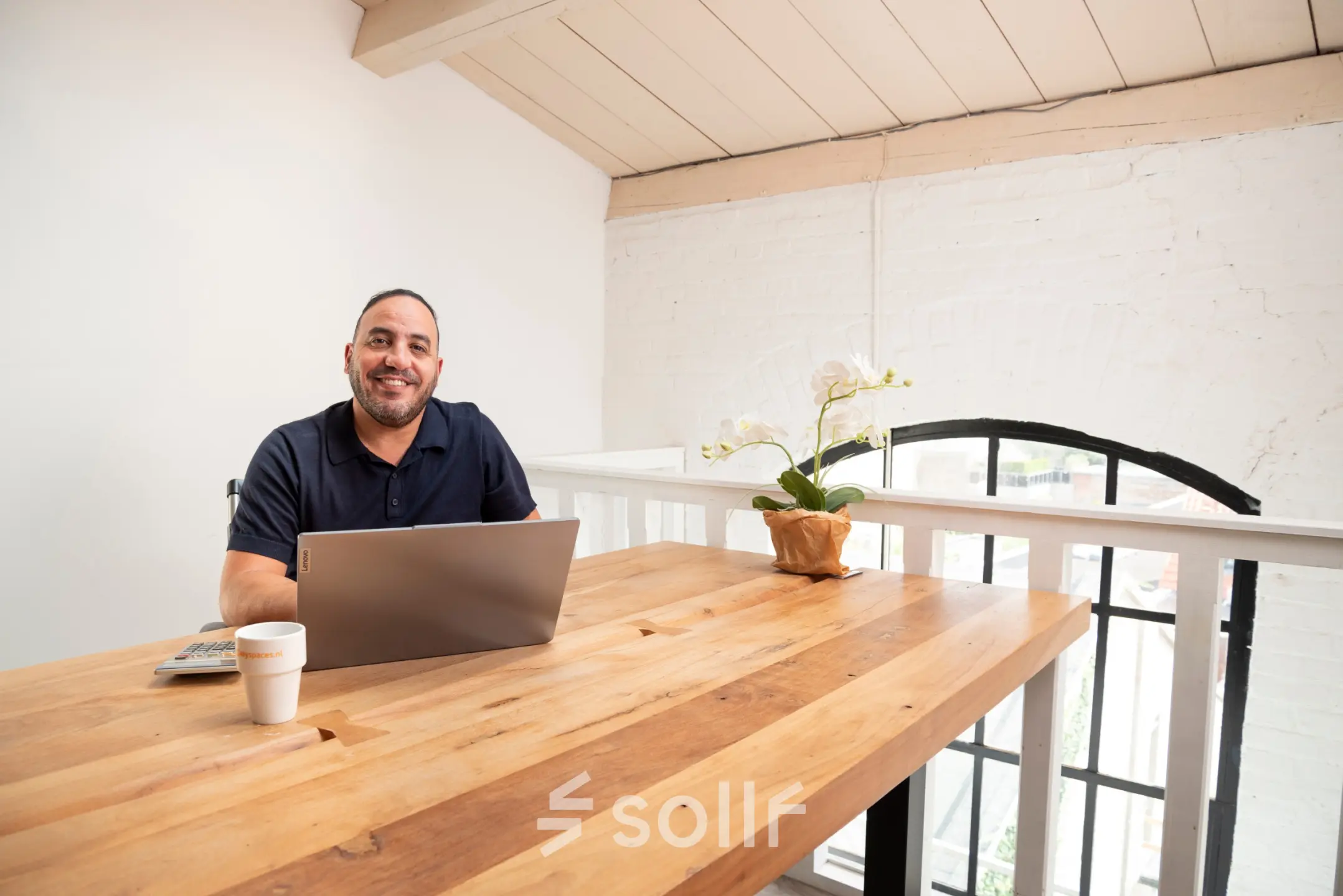 Bright office space with a wooden desk, laptop, and a person smiling while working, located at Zuidzijde Haven 39a, Bergen op Zoom. Ideal for office space rental.