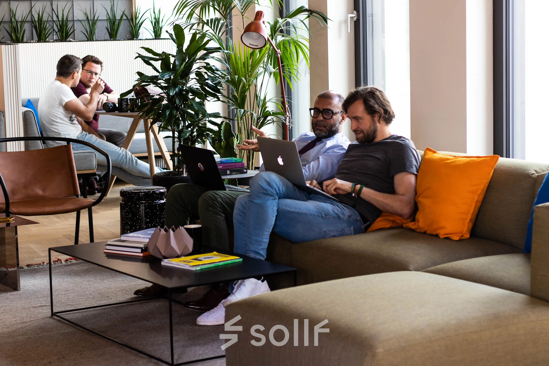 Business professionals collaborating on laptops in a modern office lounge with stylish furniture and indoor plants at Warschauer Platz 11-13, ideal for office space rental in Berlin Friedrichshain.