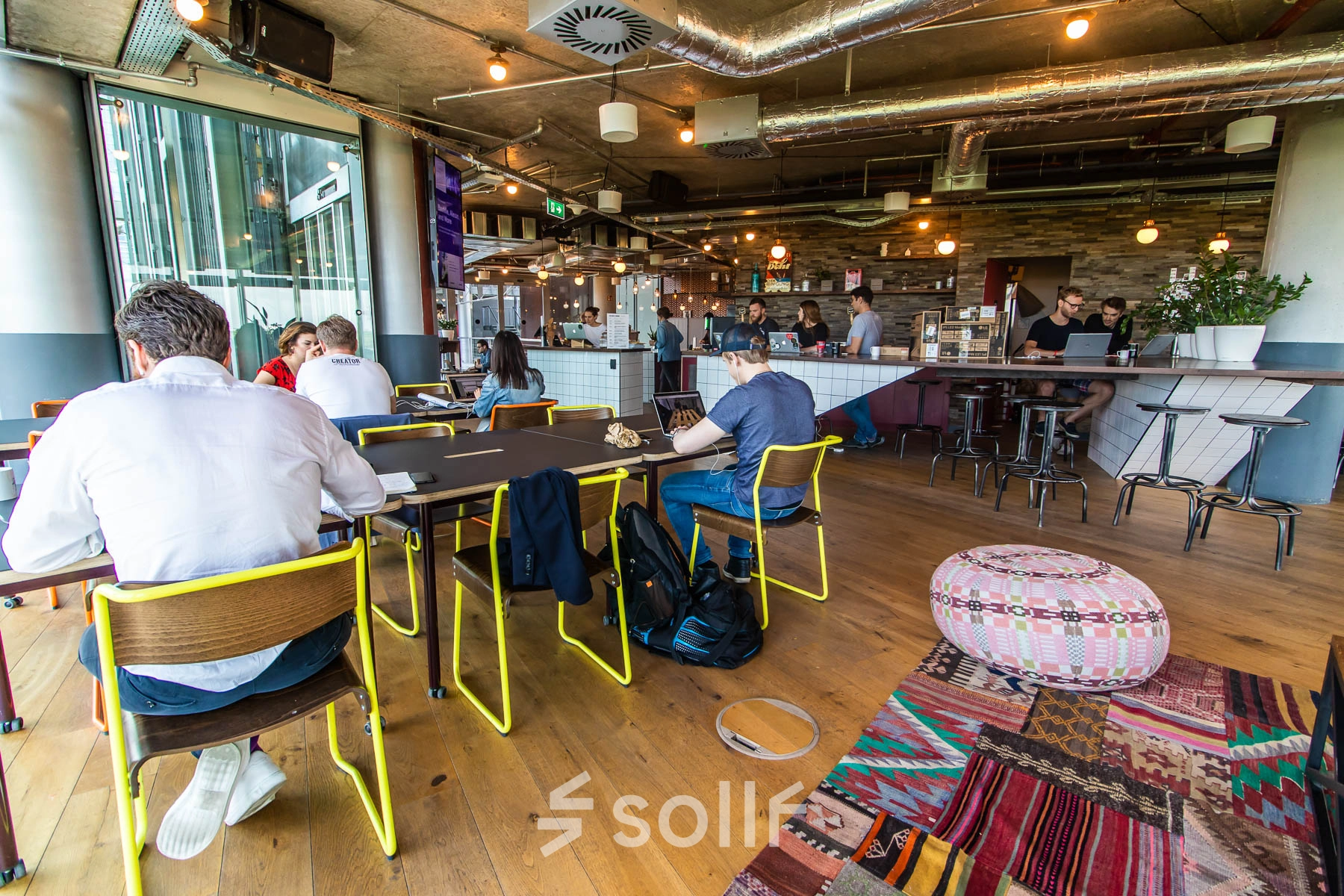 Modern office space rental at Warschauer Platz 11-13, Berlin Friedrichshain, showcasing individuals collaborating over a large table with vibrant decor and industrial lighting.