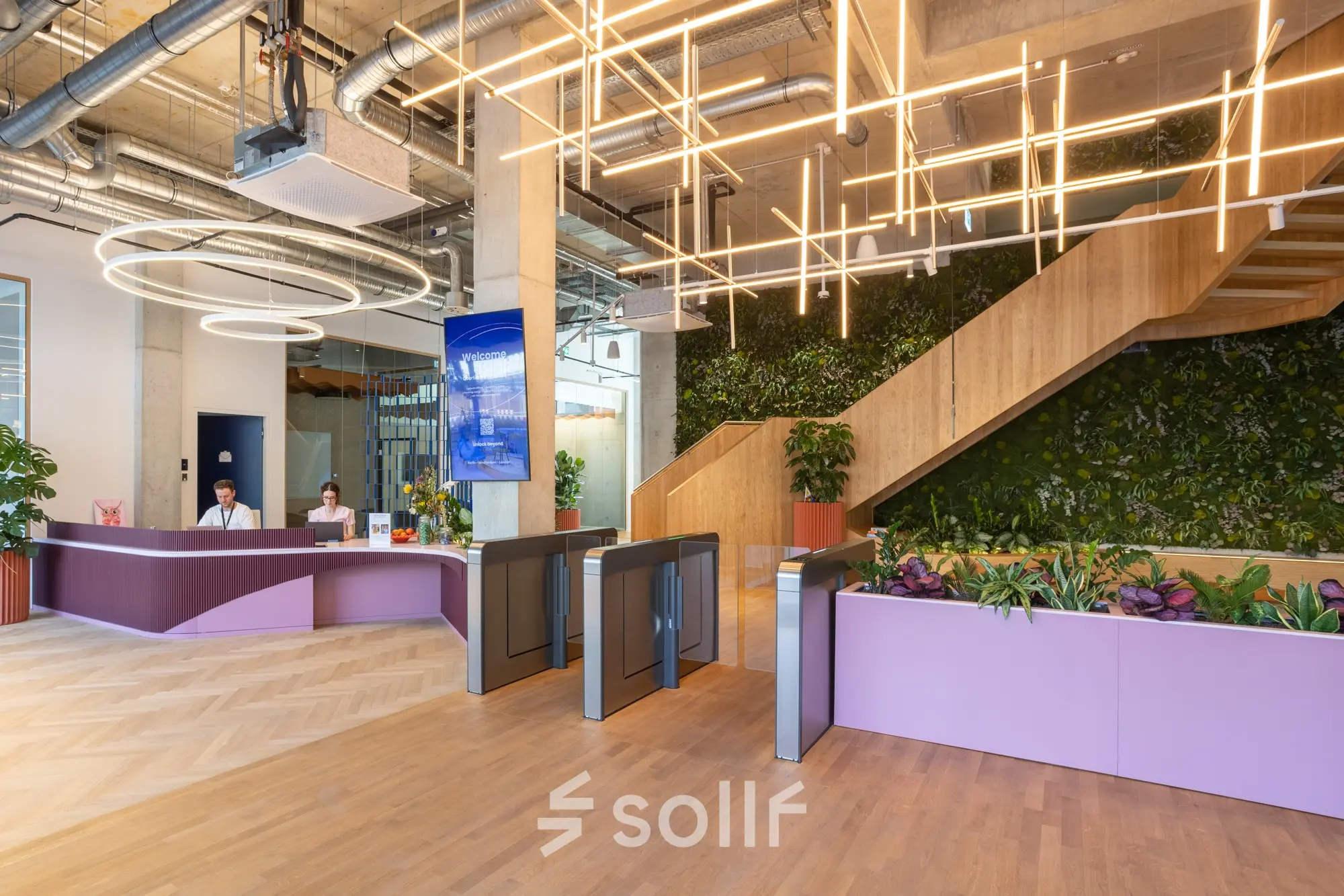 Modern reception area at an office space rental in Berlin Mitte, featuring a striking staircase, contemporary light fixtures, and a lush green wall. Two people are working at the reception desk.