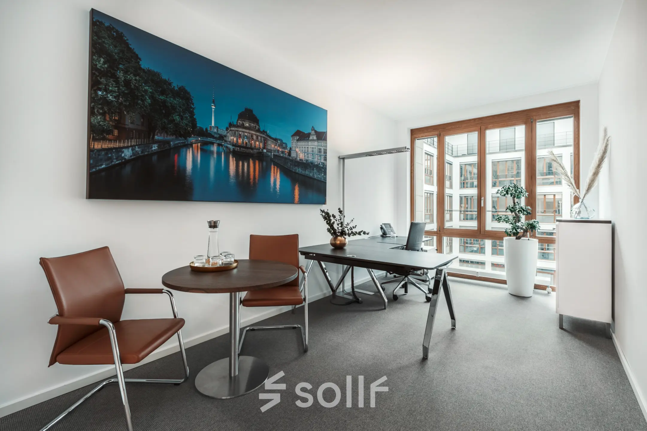 Modern Berlin Mitte office space rental at Pariser Platz 6a, featuring a meeting area with chairs, table, and work desk near large windows.