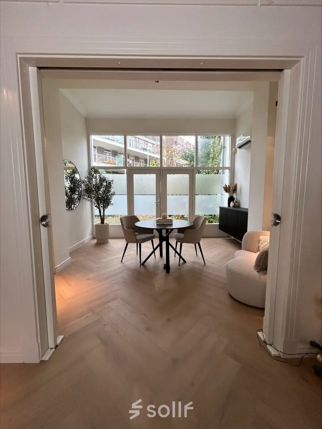 Bright office space with a round table and chairs, ideal for rent. Located in a peaceful setting with a view of the outside area through large windows.