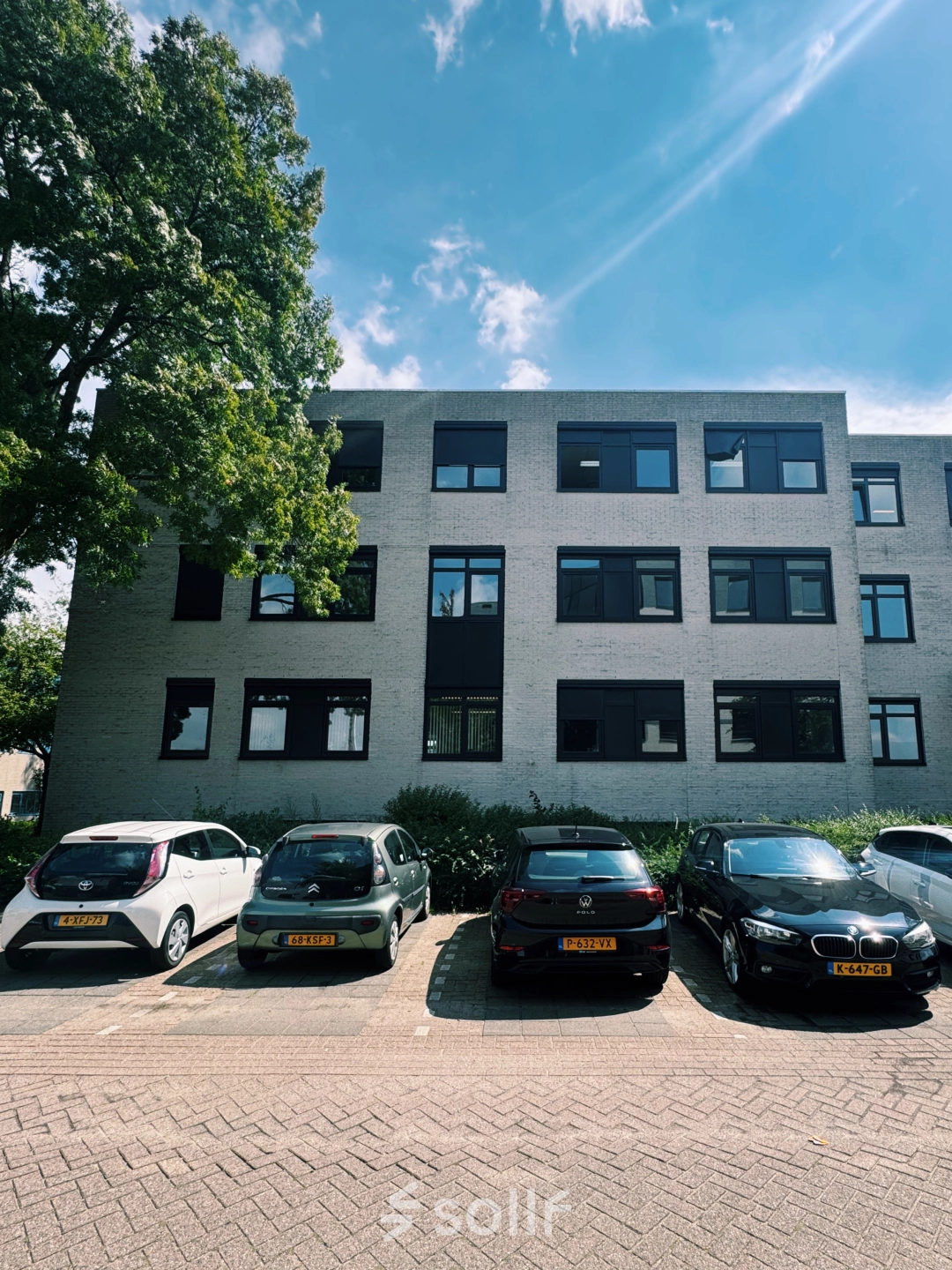 Exterior view of a modern office building at Druivenstraat 25-31 in Breda Oost, suitable for office space rental, with cars parked in front, framed by trees.