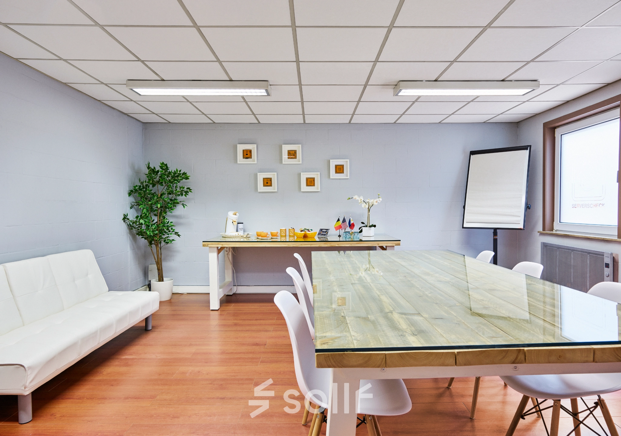 Modern office space with a long wooden table, white chairs, and a white sofa next to a refreshment area, available for rent at Leuvensesteenweg 613, Brussels Airport.