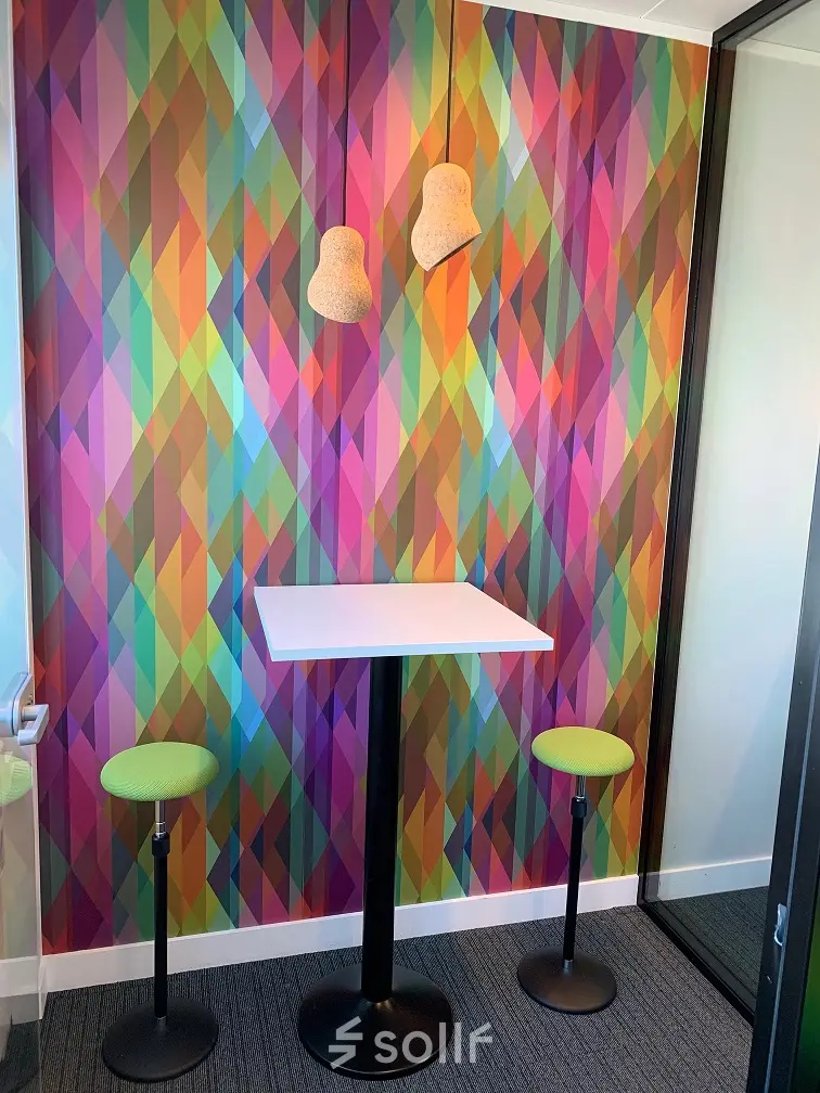Vibrant office corner at Rue Joseph Stevens 7, Brussels Center, featuring colorful geometric walls with a white table and green stools, perfect for office space rental.