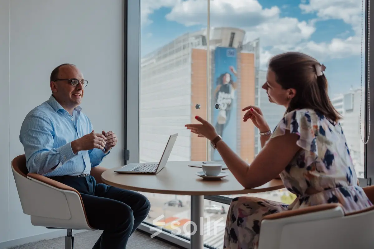 Two business professionals engage in a discussion at a round table in a modern office with a view of Brussels European District, ideal for office space rental.