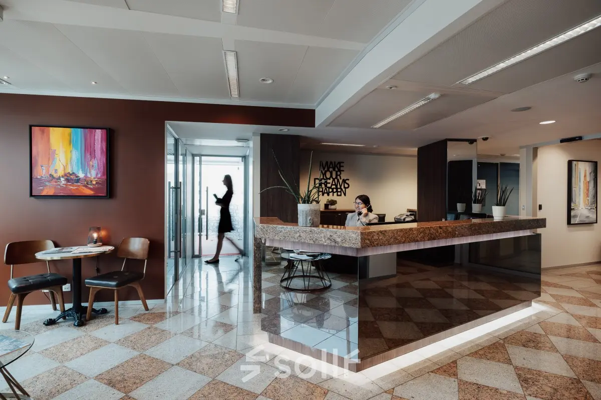 Modern office with a reception desk and a small meeting area. A person works at the desk while another walks by, set in Brussels European District.