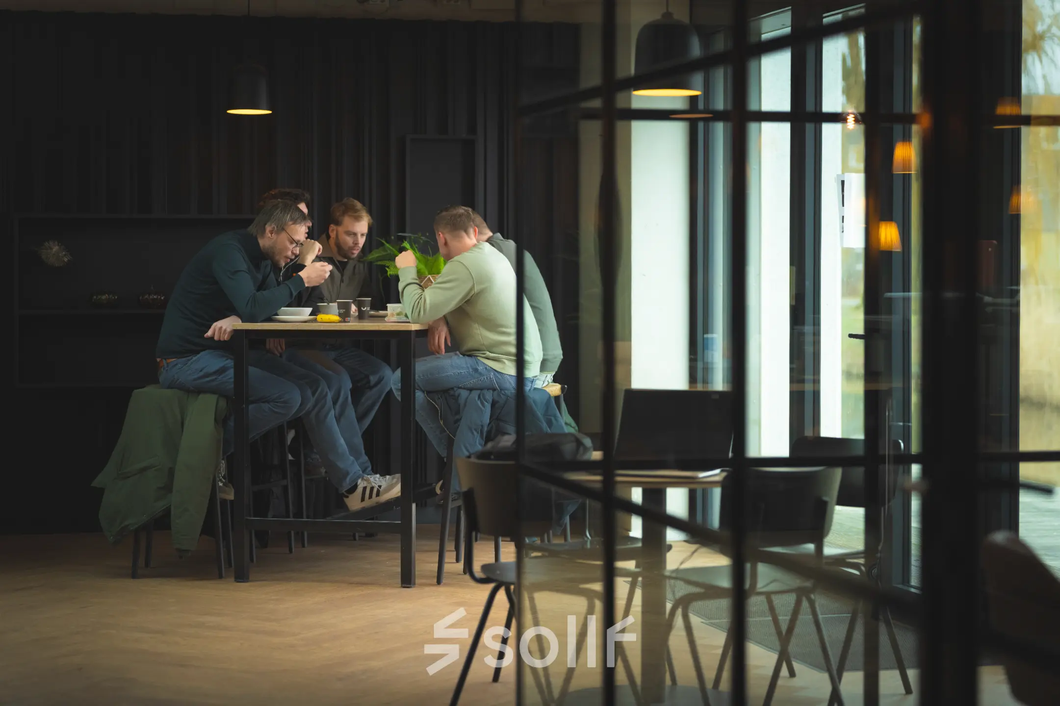 Three people sit at a tall table in a modern office space at Rietbaan 2-12, enjoying a meal while discussing business. The room is stylishly furnished with large windows.