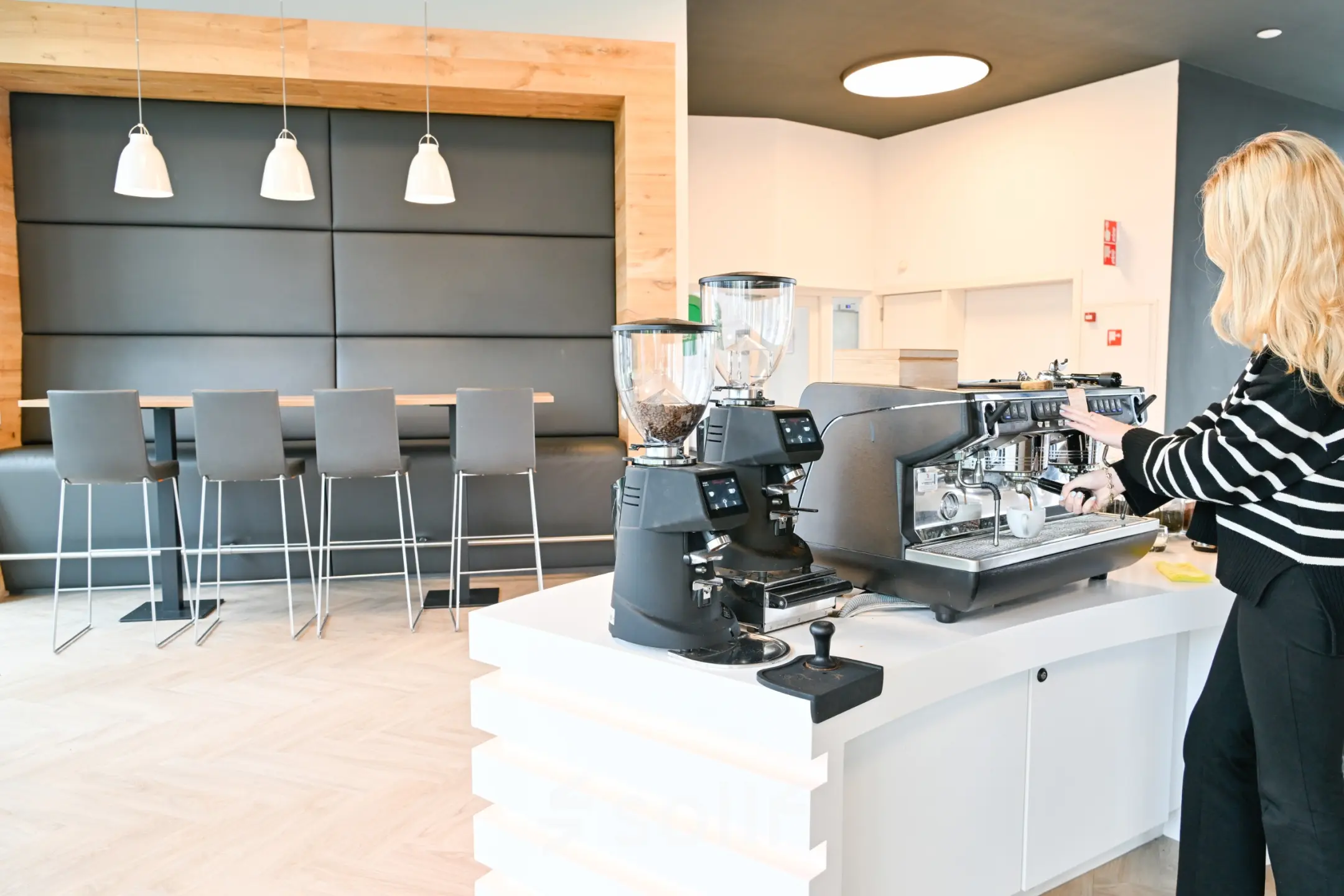 A bright kitchen area with a person making coffee, featuring modern bar stools and light wood accents, ideal for office space rental.
