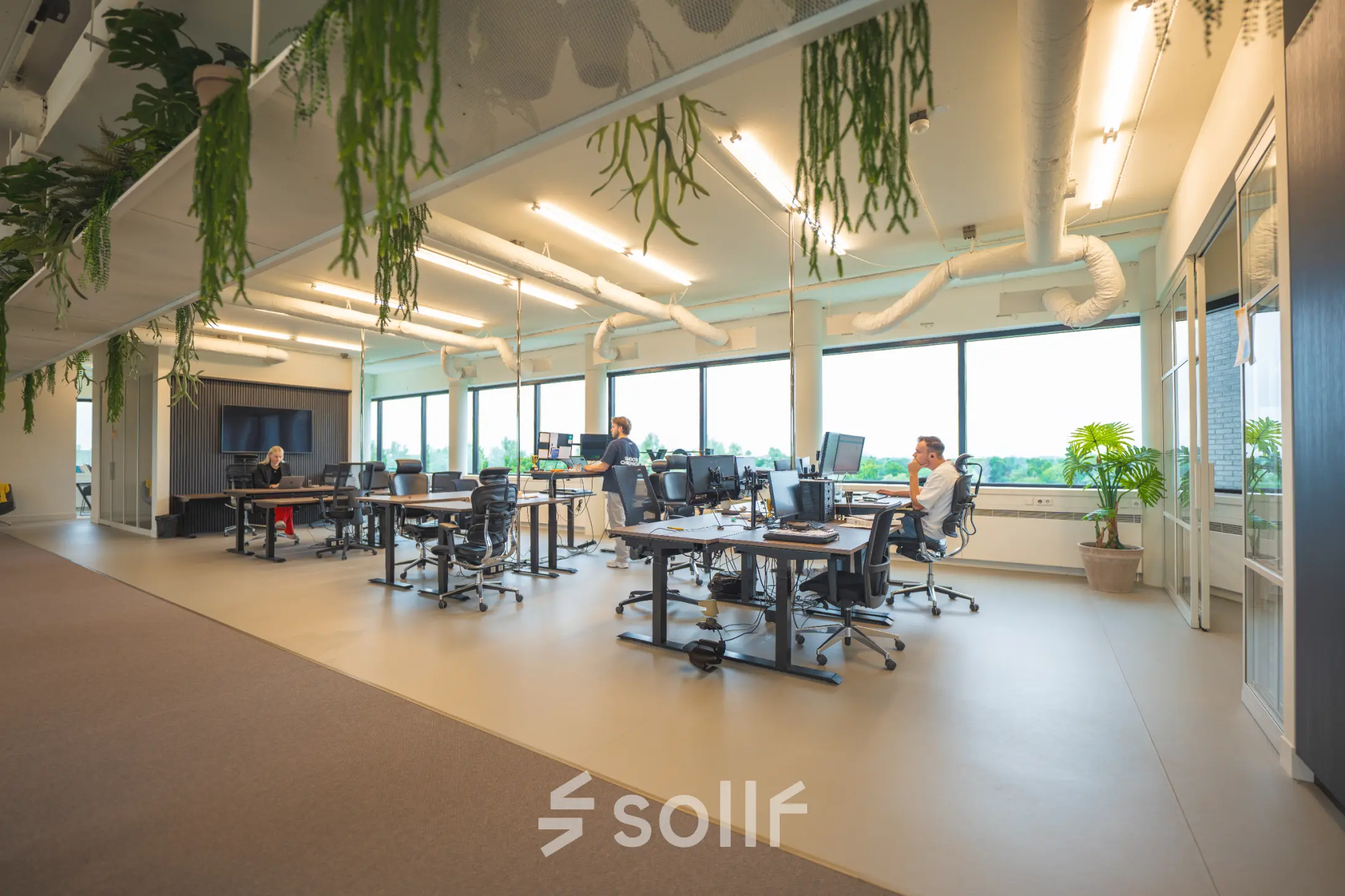 Modern office space with multiple desks, computers, and people working at Hambakenwetering 1 in Den Bosch. Ideal for those looking to rent office space with a vibrant atmosphere and ample natural light.